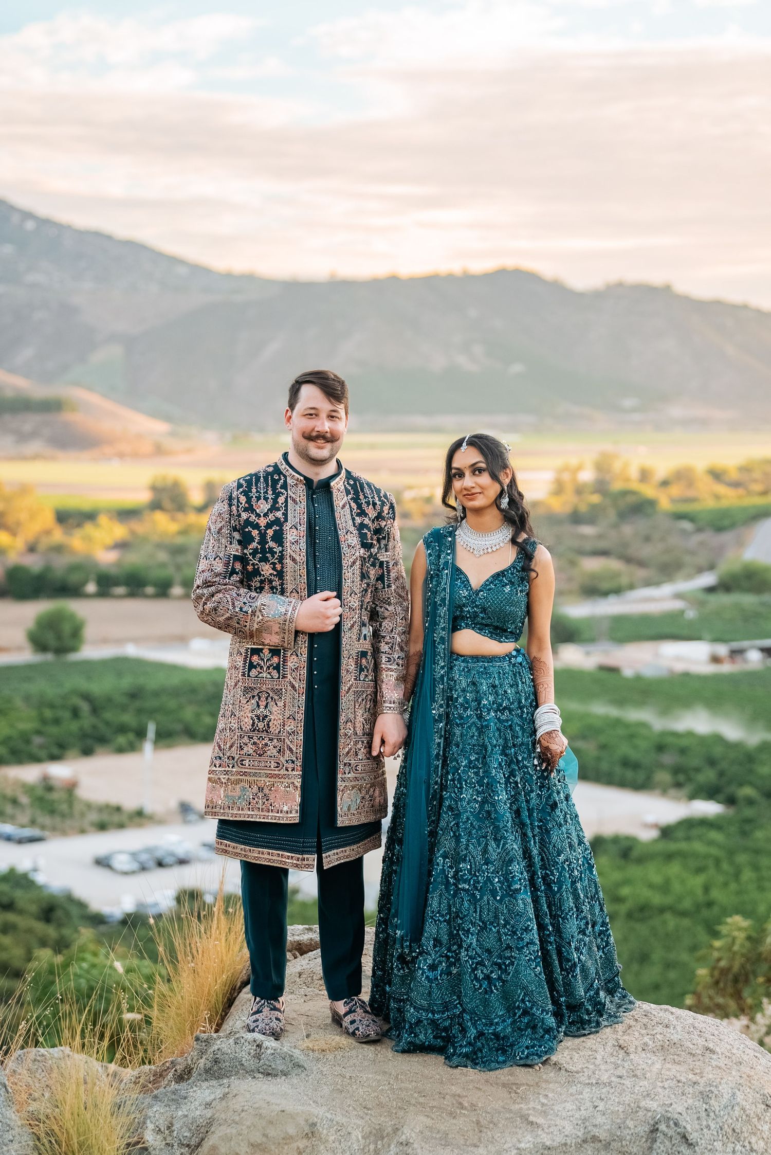 Couple in traditional Indian wedding attire standing on rocks with mountain valley backdrop at sunset.