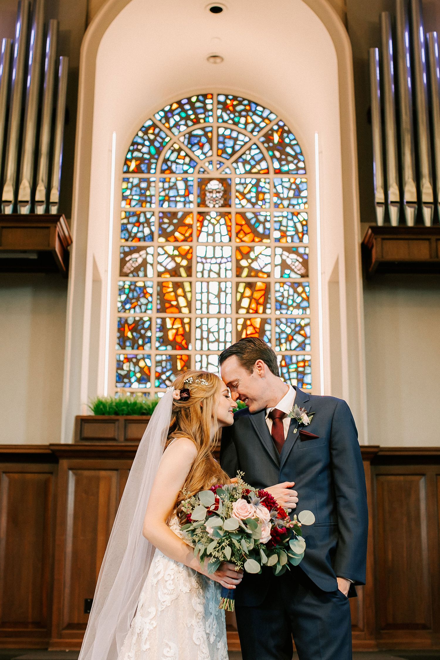 Bride and groom share a romantic moment in front of a stunning stained glass church window on their wedding day.