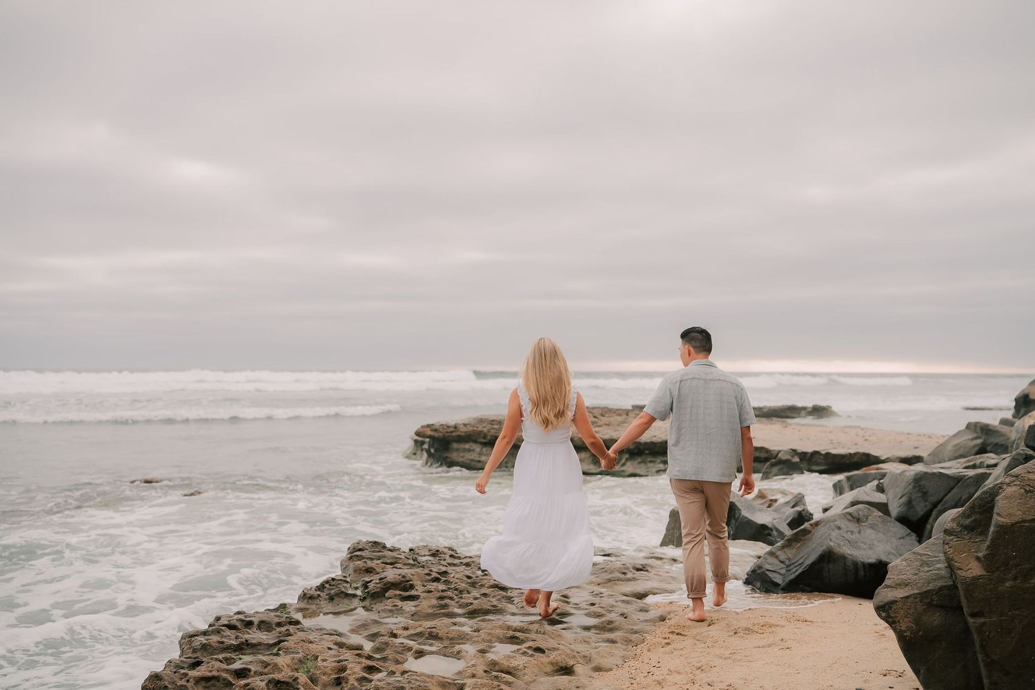 Couple holding hands walking along rocky beach shoreline with ocean waves and overcast sky.