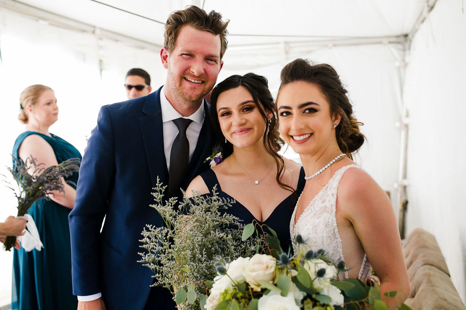 Bride holding bouquet poses with groom in navy suit and guest at outdoor wedding celebration.