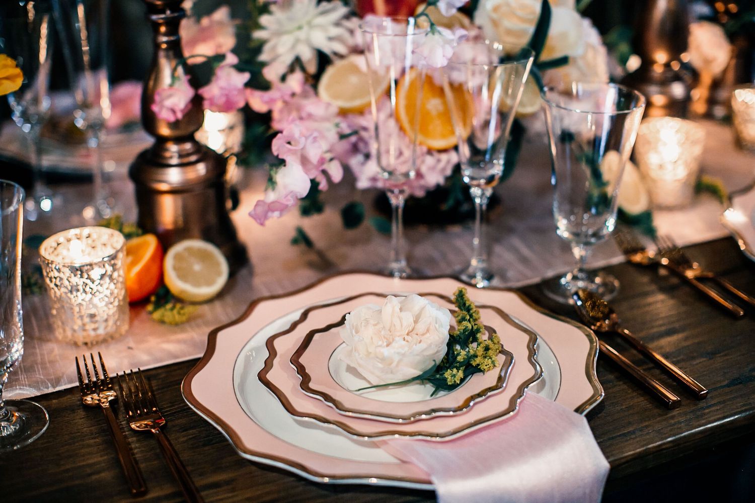 Elegant wedding table setting with gold cutlery, white floral plates, pink napkin, and lush floral centerpiece.