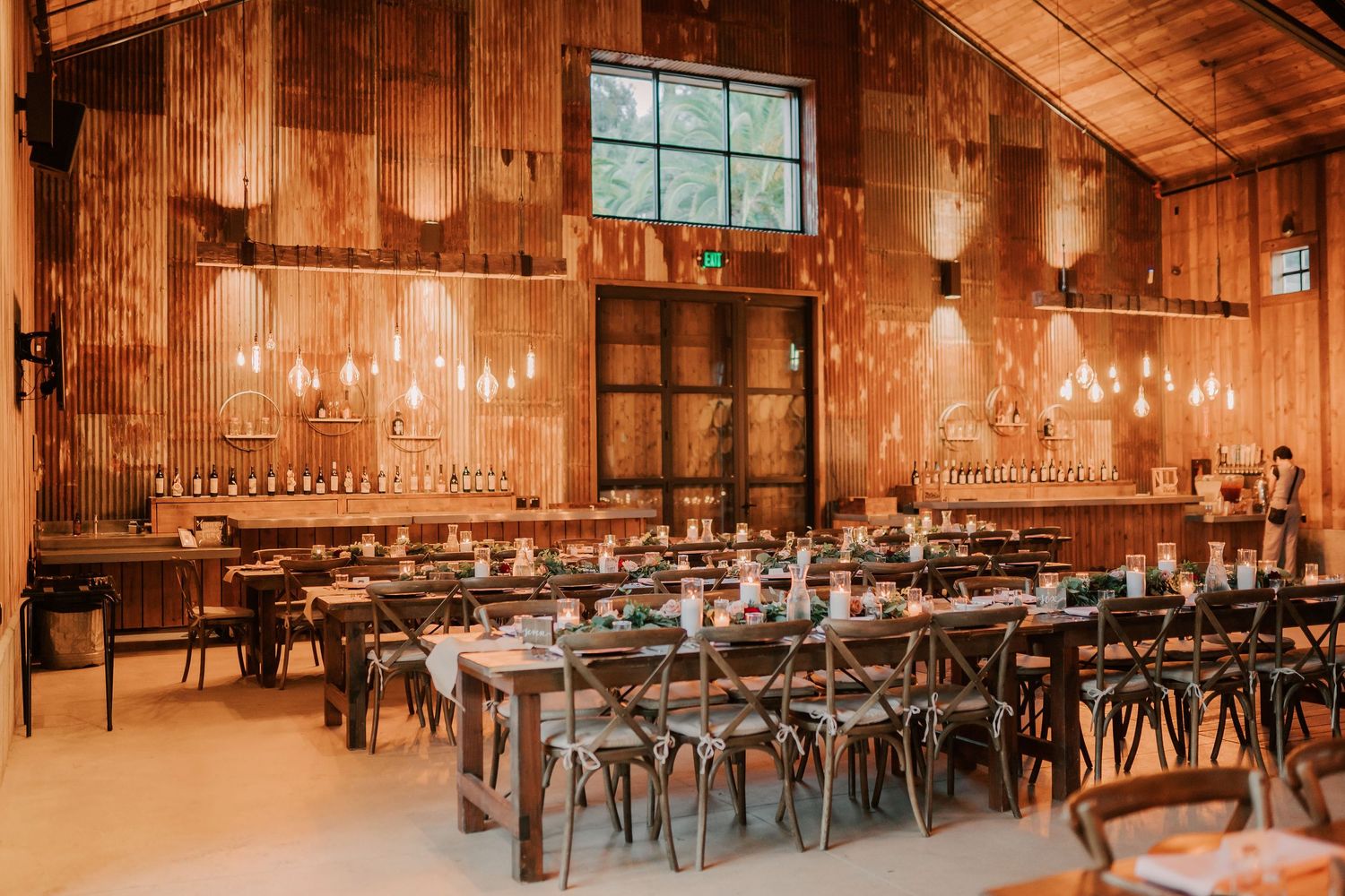 Rustic barn wedding reception with wooden walls, string lights, long dining tables, and cross-back chairs.