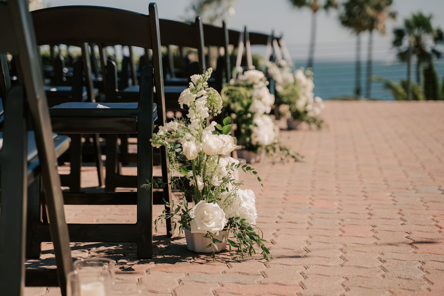 White floral arrangements line the aisle of an outdoor oceanfront wedding ceremony with black chairs and a brick patio.