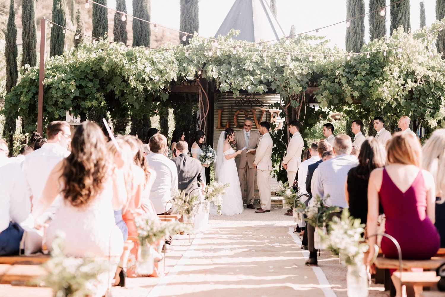 Outdoor wedding ceremony with guests seated along a flower-lined aisle beneath a lush green arbor.
