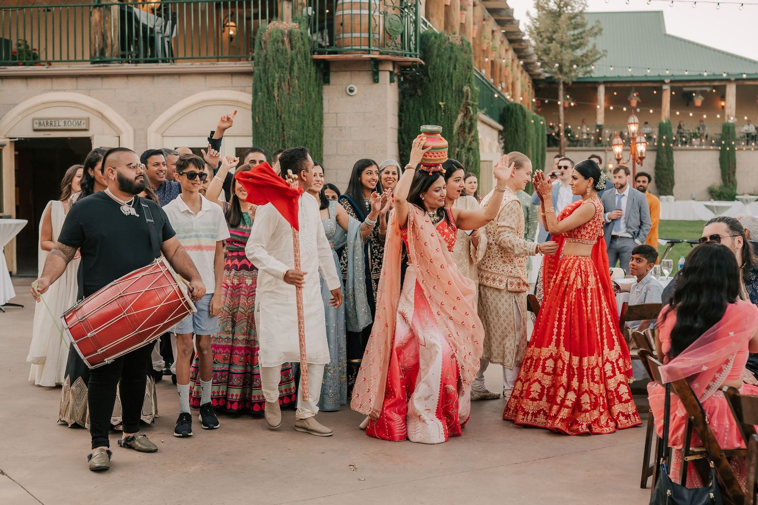 Bride in red lehenga celebrates with guests and dhol drummer during festive Indian wedding baraat.