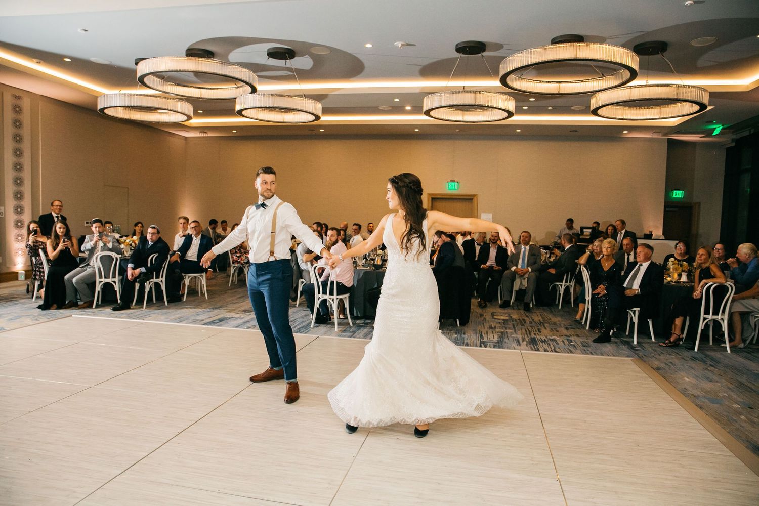 Bride and groom share first dance on white dance floor surrounded by seated wedding guests under circular chandeliers.