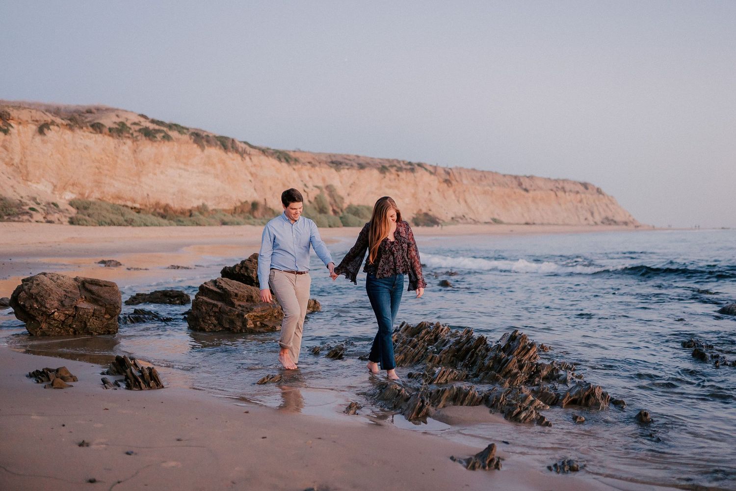 Couple holding hands walking on a rocky beach at sunset with golden cliffs in the background.