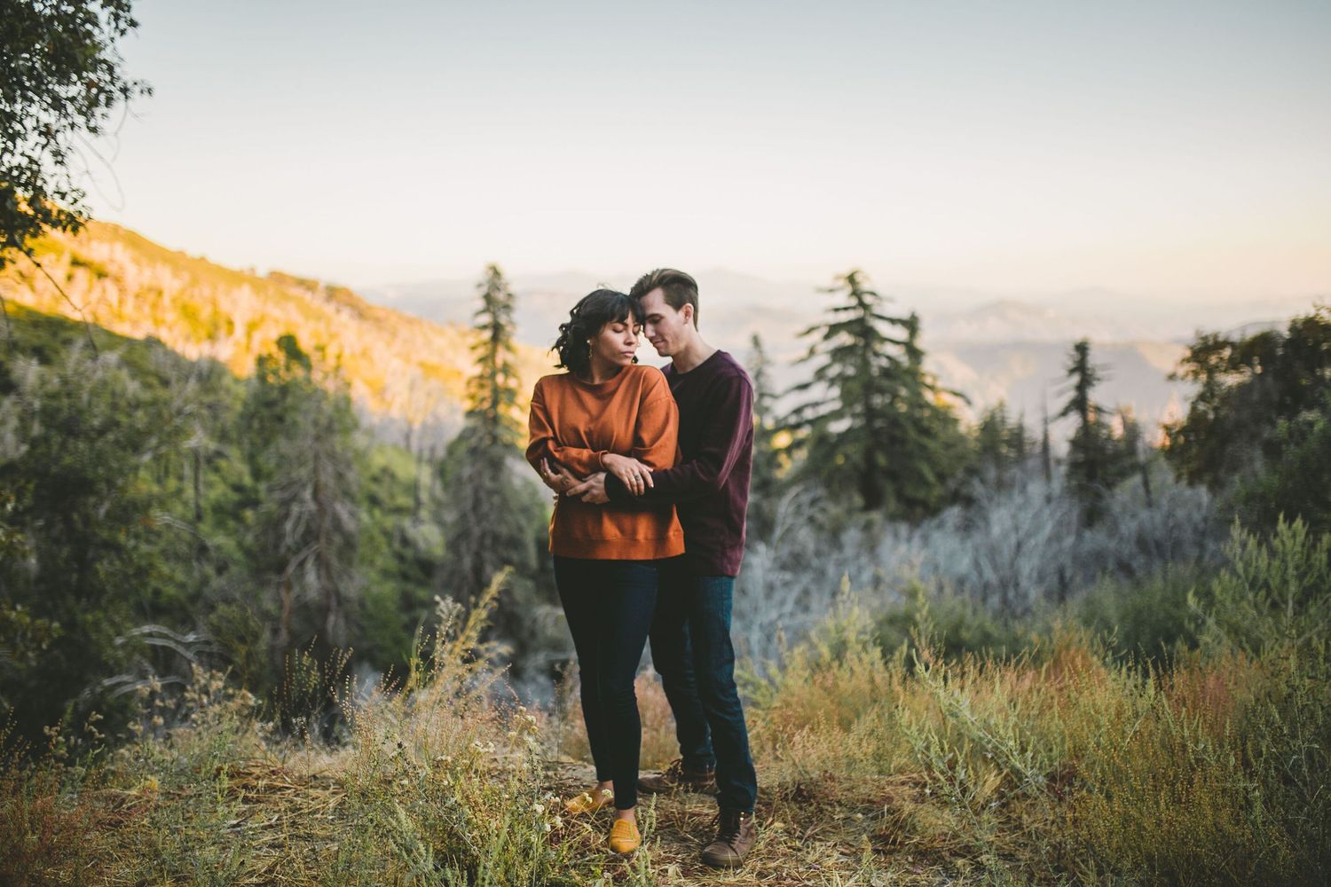 Couple embracing at golden hour on a mountain overlook surrounded by pine trees and autumn foliage.