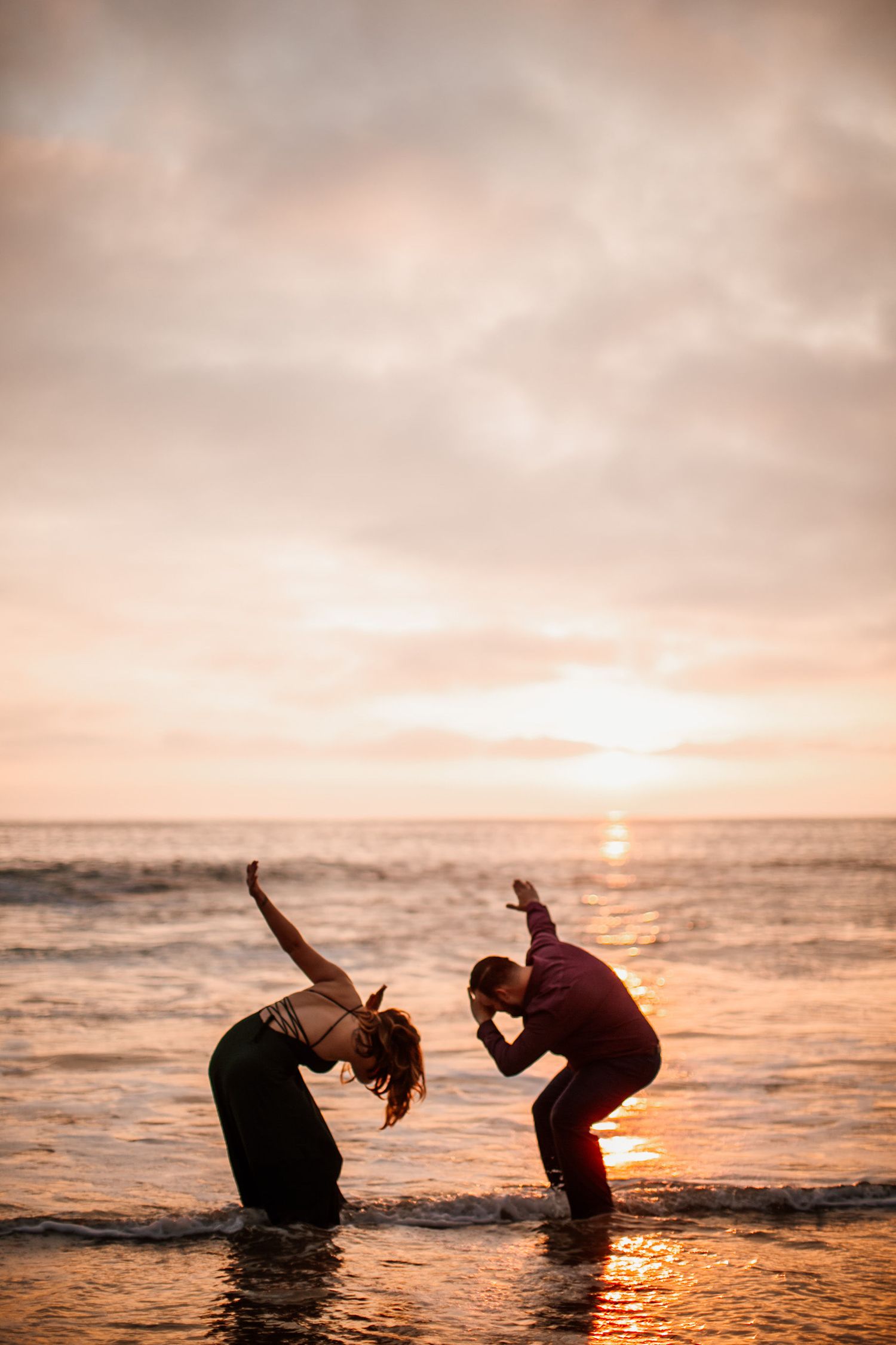 Two people practicing yoga poses in ocean waves at sunset, silhouetted against a golden sky.