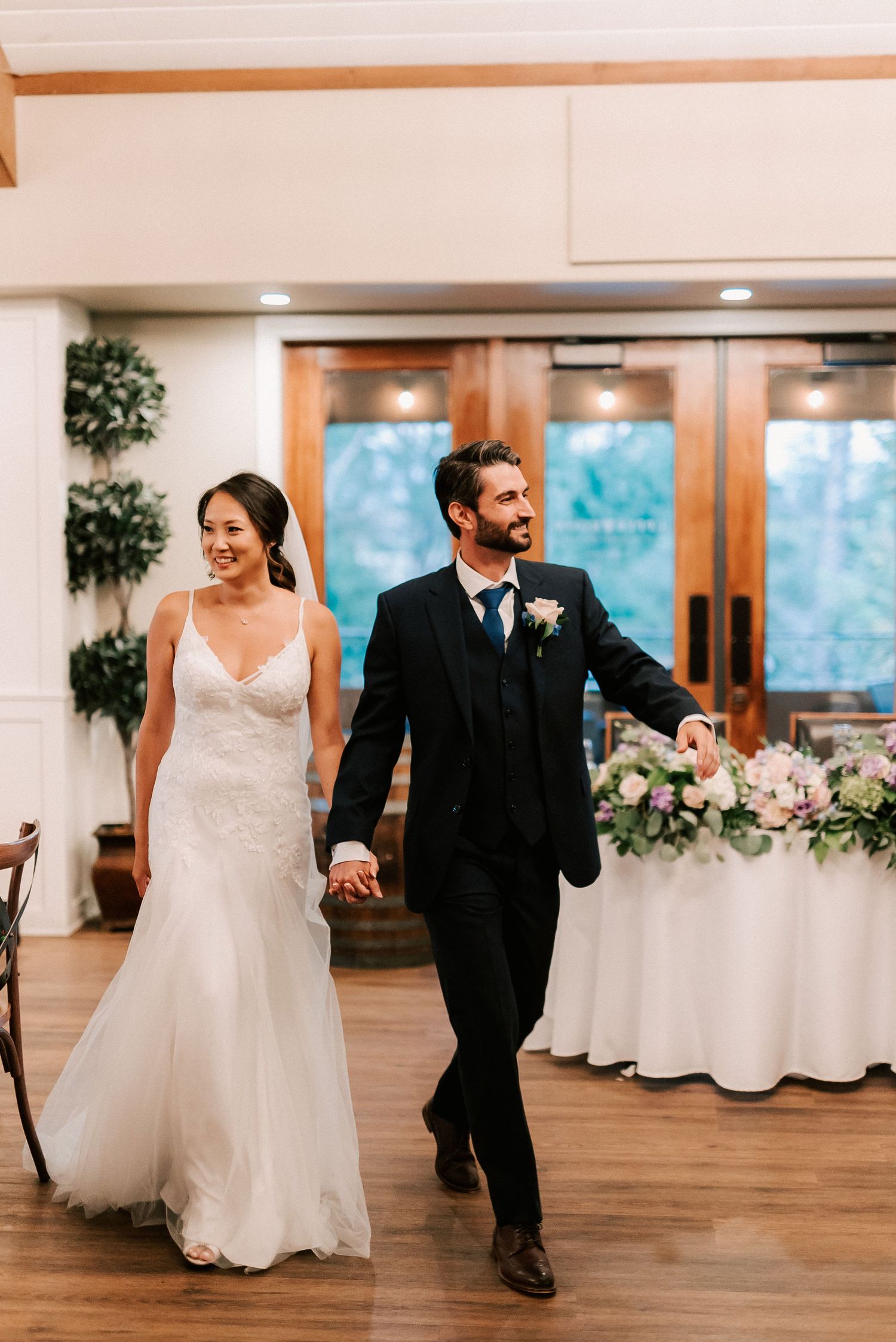 Bride in lace gown and groom in black suit hold hands while making their grand entrance at elegant wedding reception.