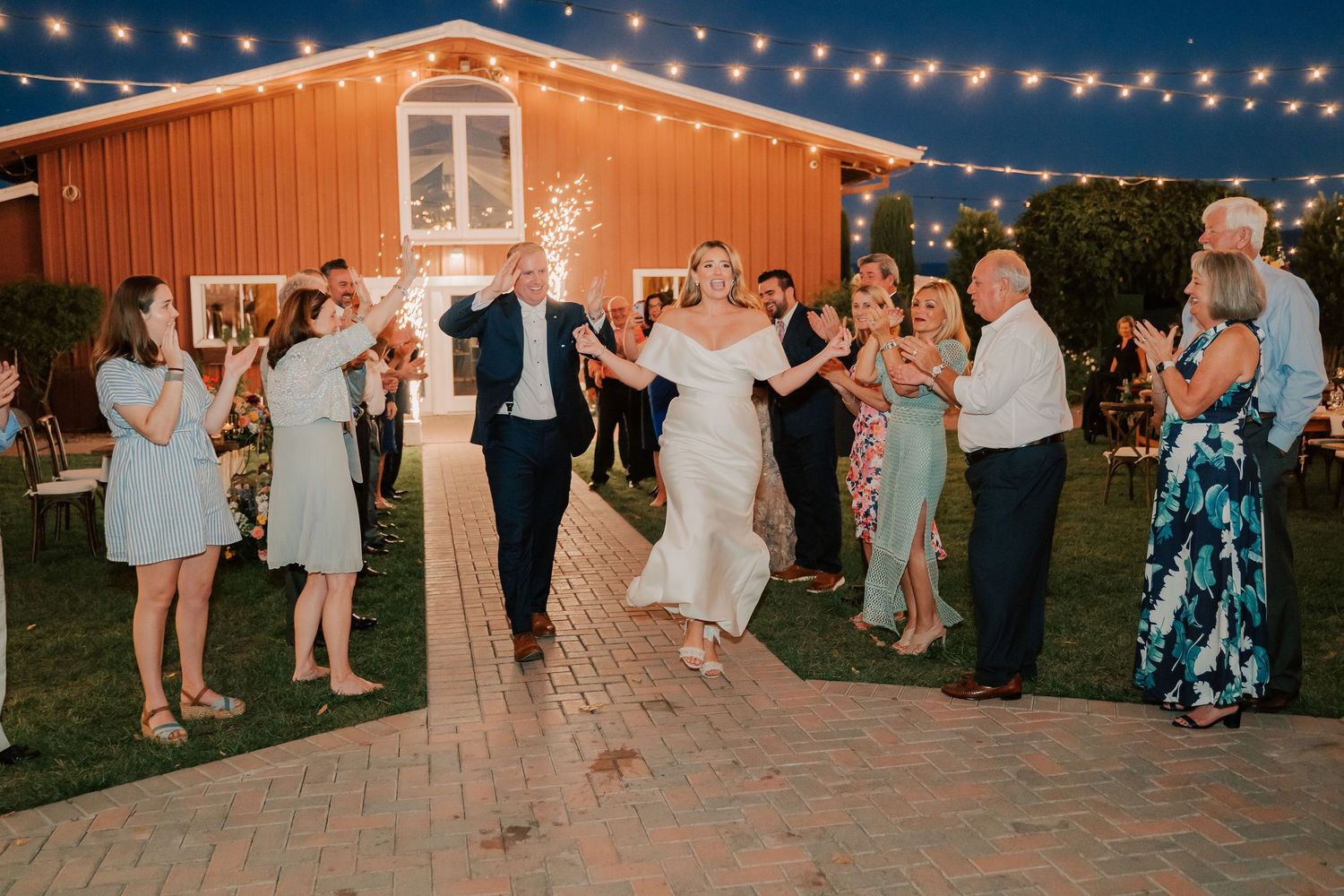 Bride and groom exit wedding reception with sparklers outside a red barn venue at night.
