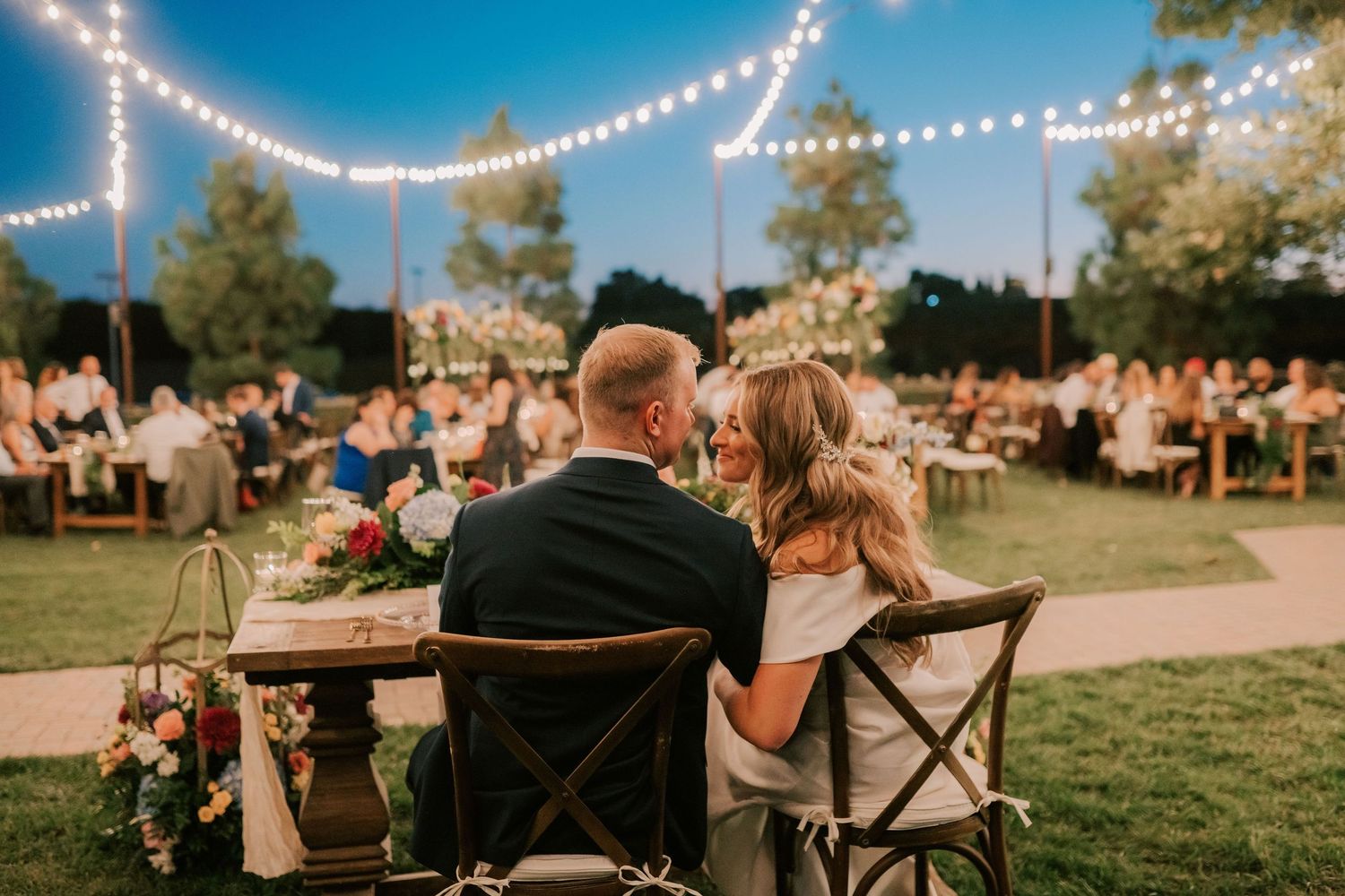 Newlywed couple seated at sweetheart table during outdoor evening wedding reception with string lights.