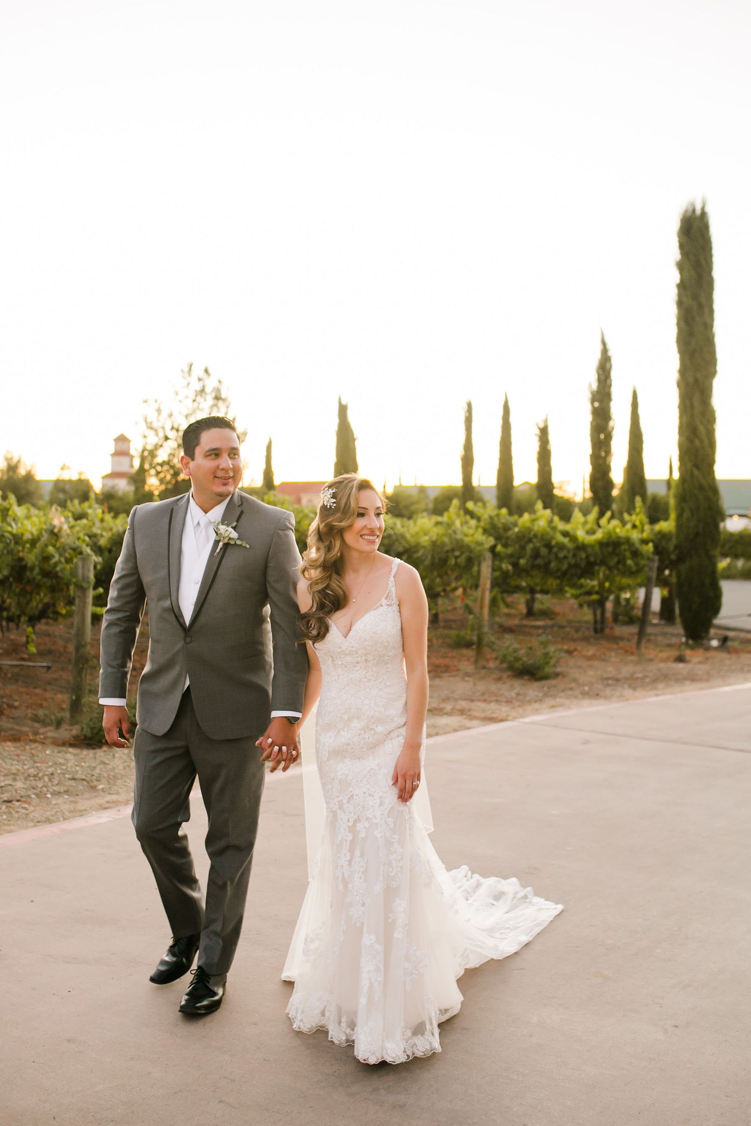 A bride and groom walk hand-in-hand through a vineyard at golden hour, she in a lace gown, he in a gray suit.