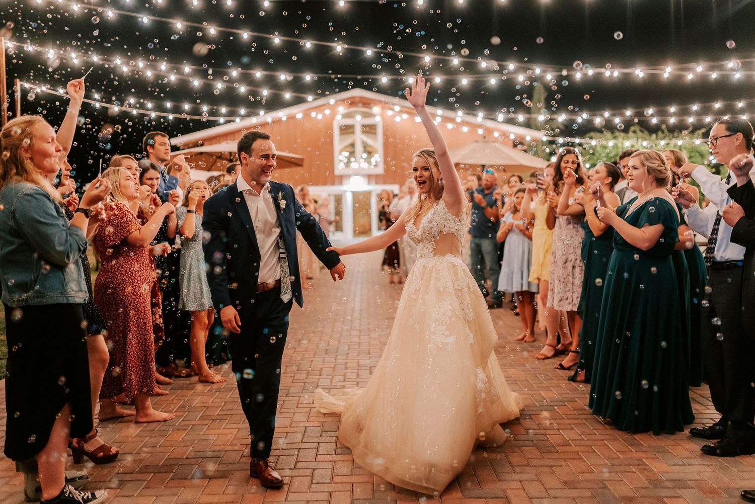 Bride and groom exit wedding reception under string lights with confetti, surrounded by cheering guests.