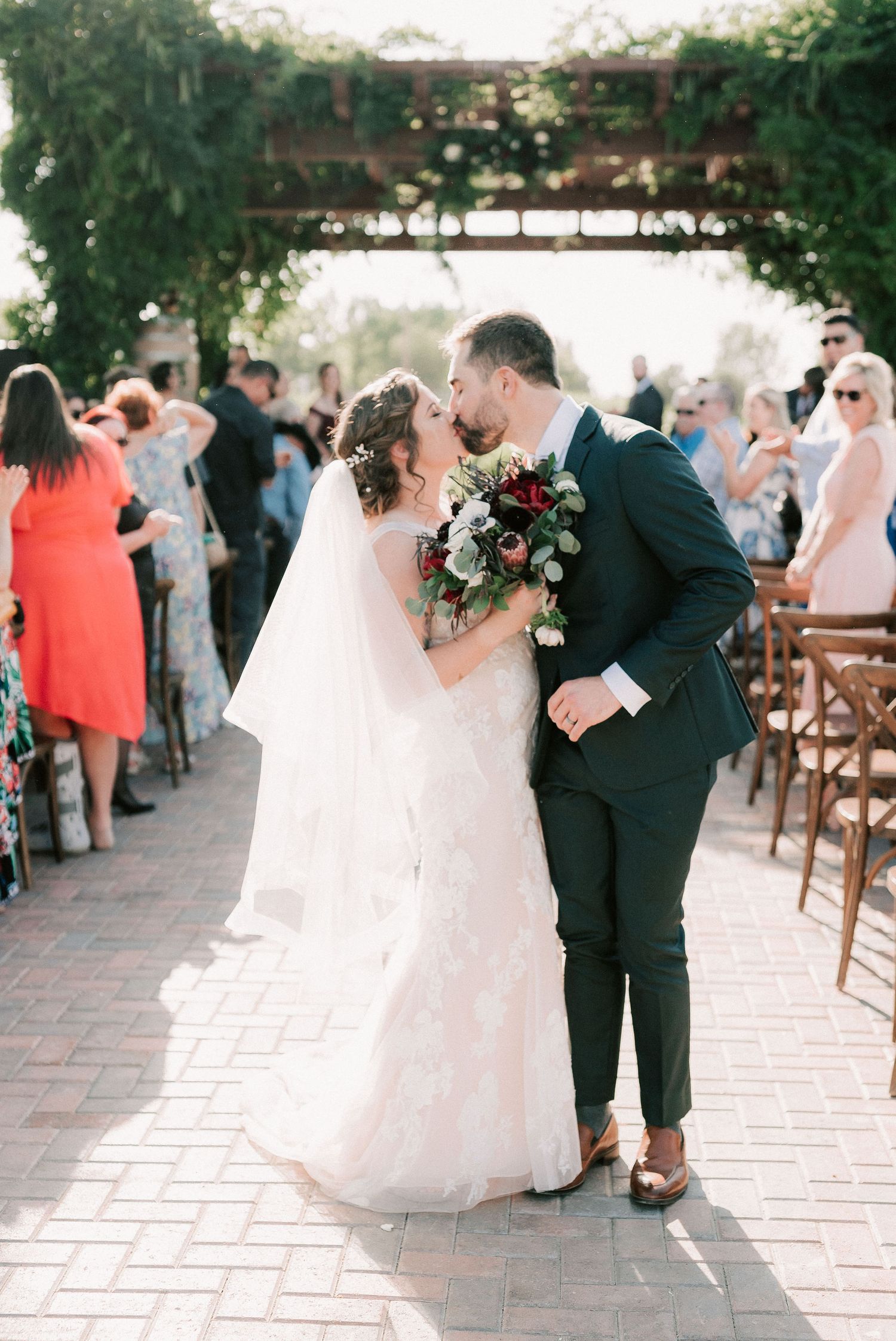 Bride and groom share a first kiss as newlyweds at an outdoor wedding ceremony with guests applauding.