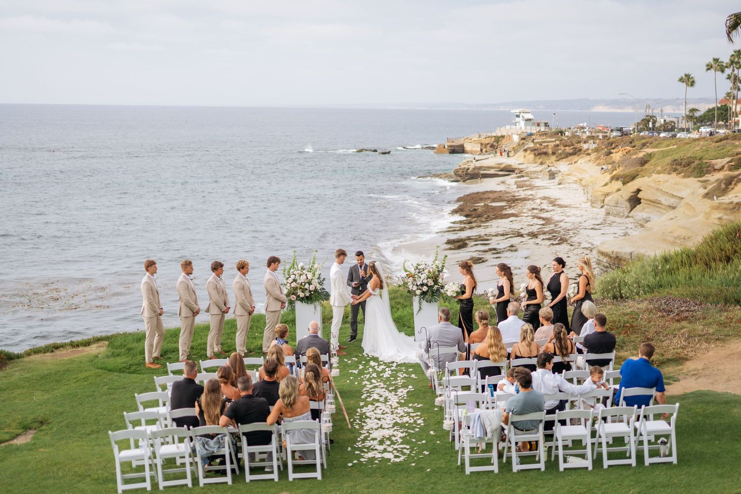 Outdoor wedding ceremony on coastal cliffside with white chairs, bridal party, and ocean backdrop in La Jolla, California.