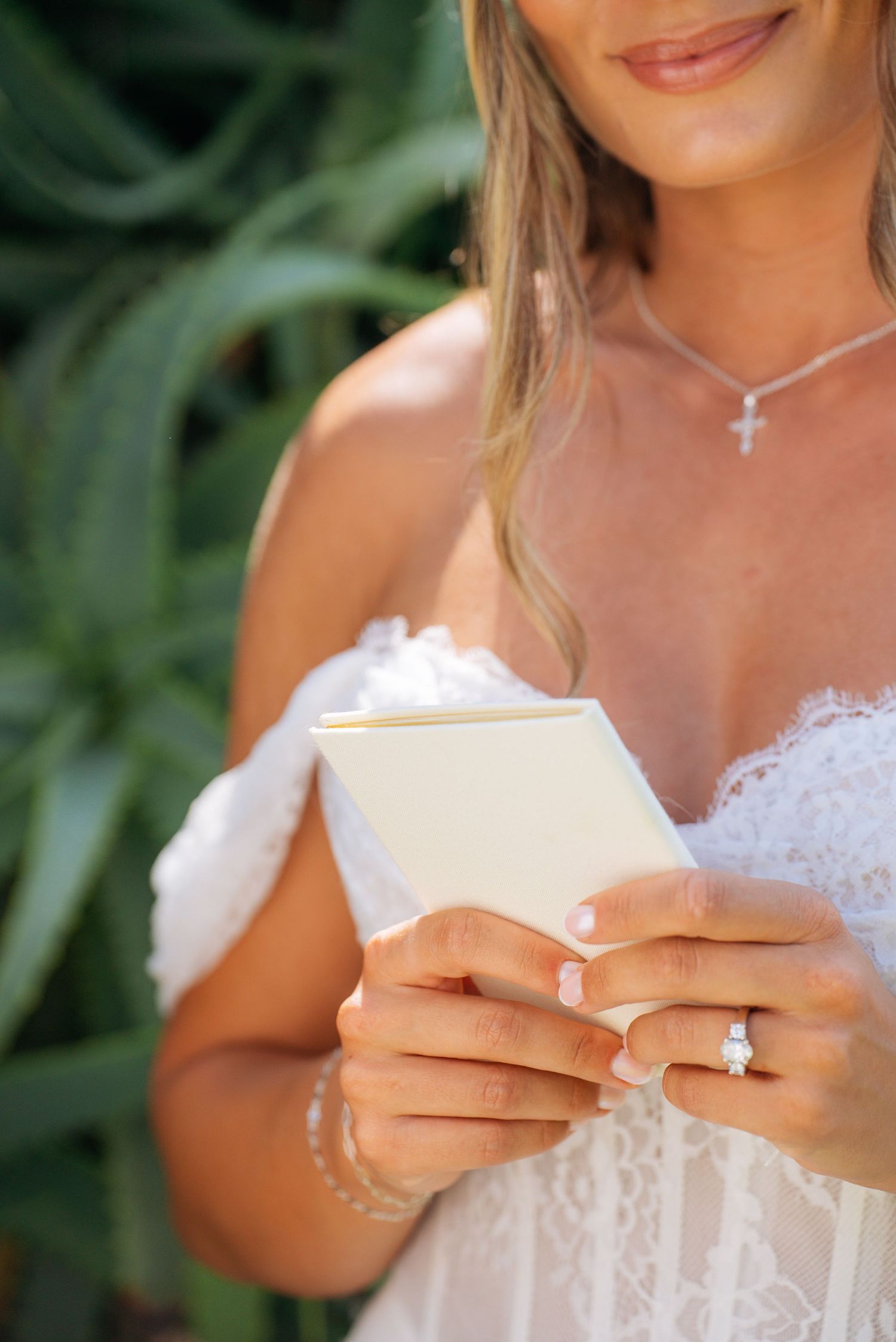 Bride in lace gown reads vow card, wearing diamond ring and cross necklace, green foliage background.