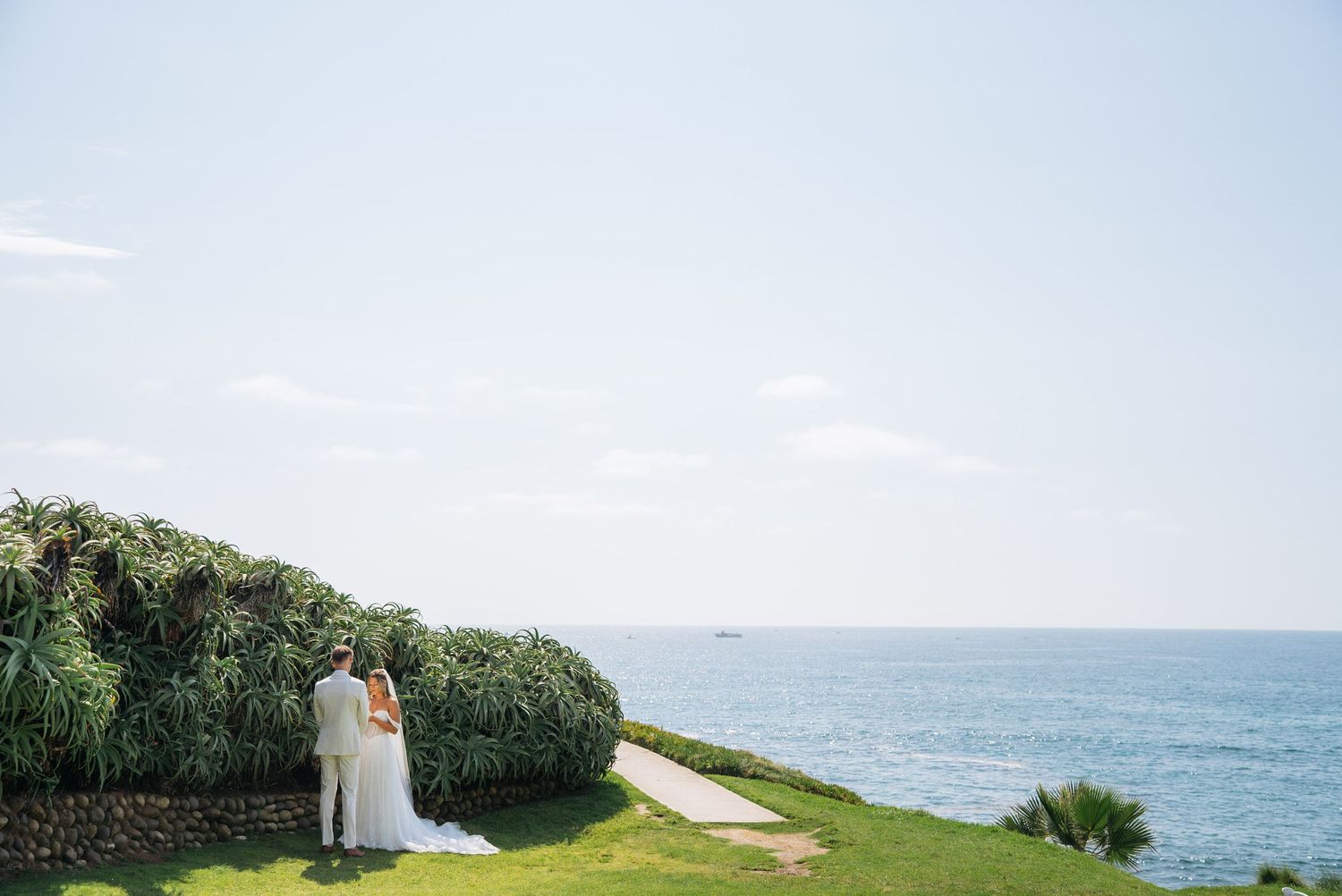 Bride and groom stand together on lush green cliffside overlooking the ocean on their wedding day.