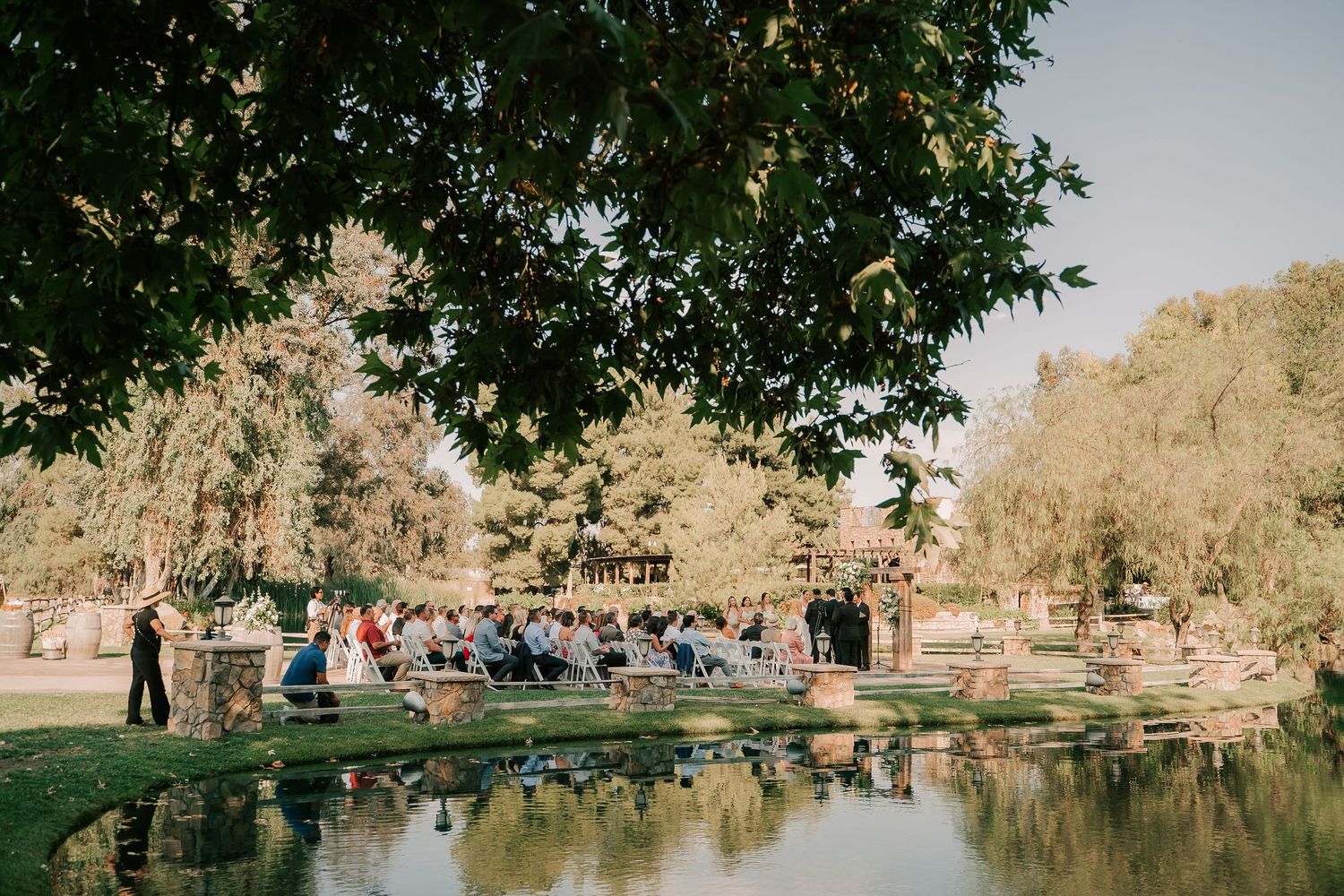 Outdoor wedding ceremony beside a tranquil pond surrounded by lush green trees and landscaped gardens.