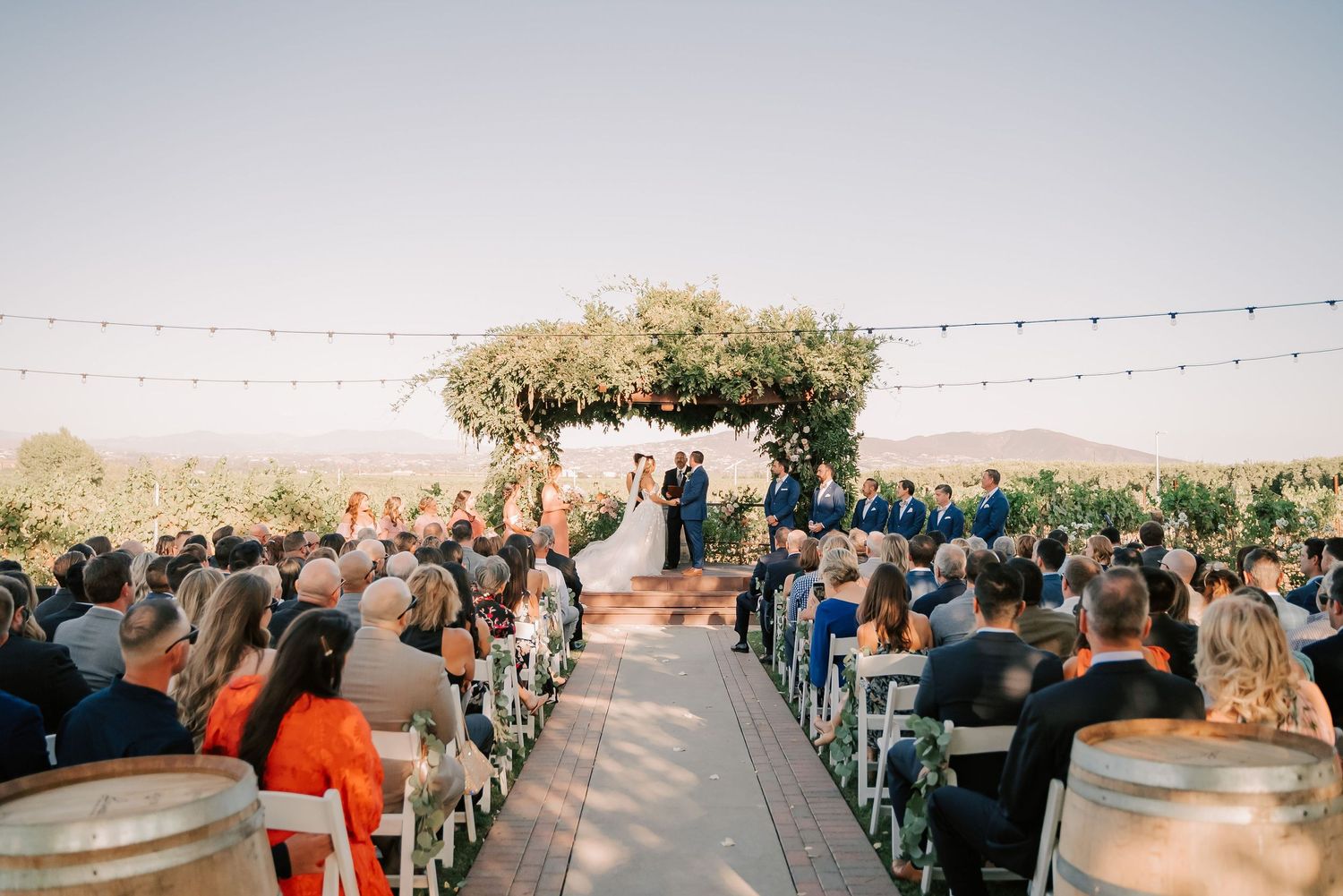 Outdoor wedding ceremony with guests seated in white chairs, string lights overhead, and couple at floral altar with vineyard views.