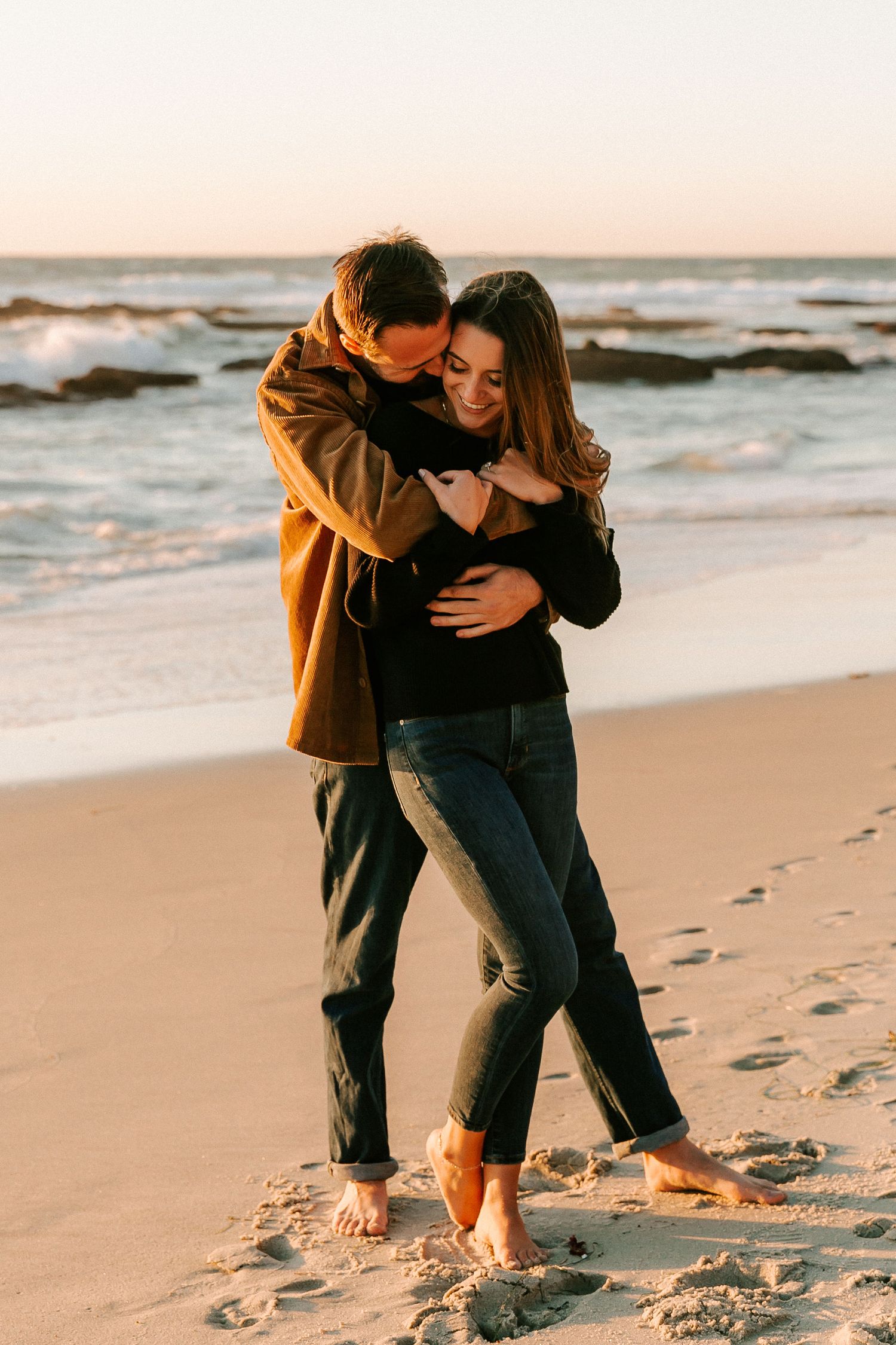 Couple embracing on sandy beach at golden hour sunset, man hugging woman from behind.