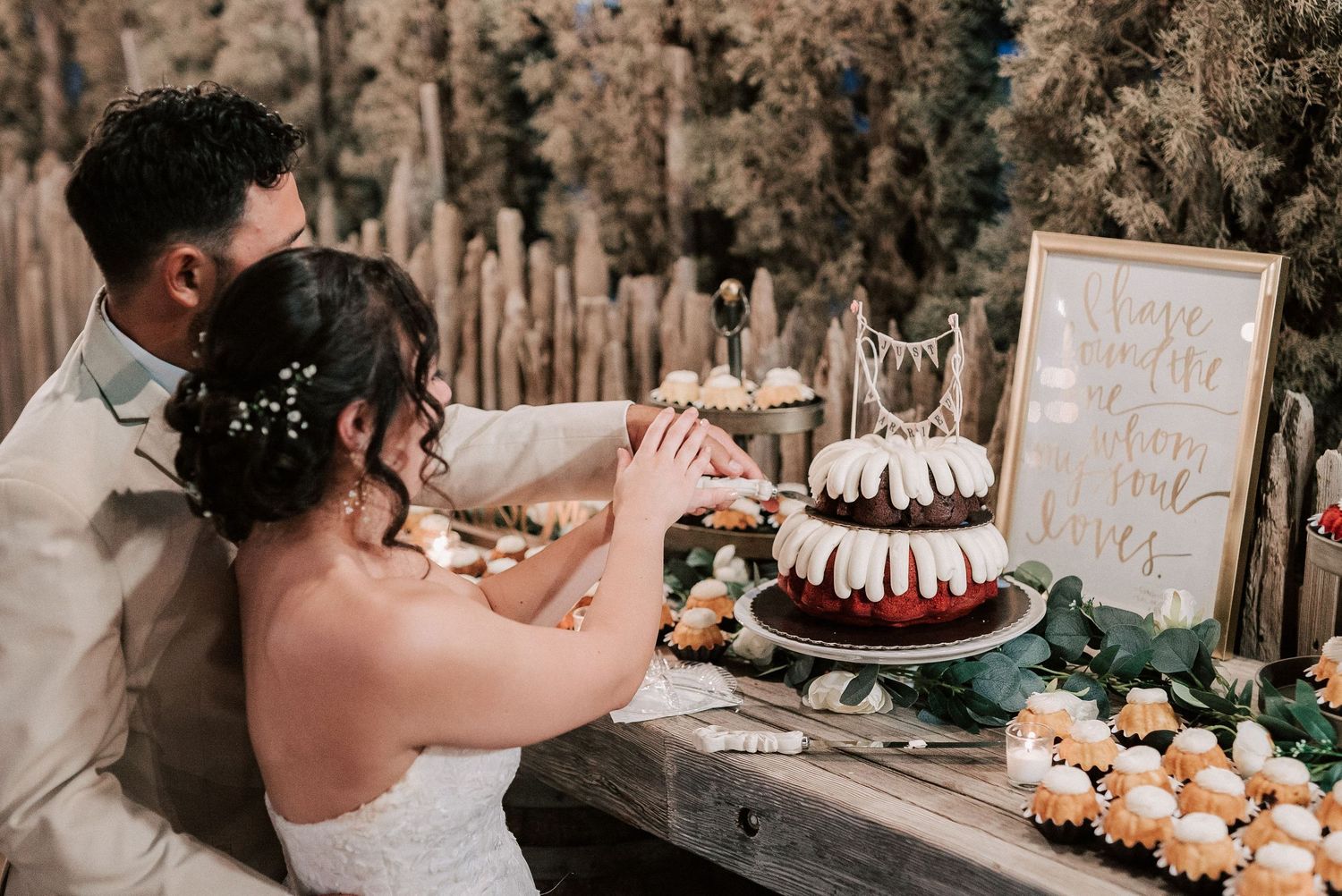 Bride and groom cutting a rustic wedding cake at an outdoor dessert table with flowers and a decorative sign.