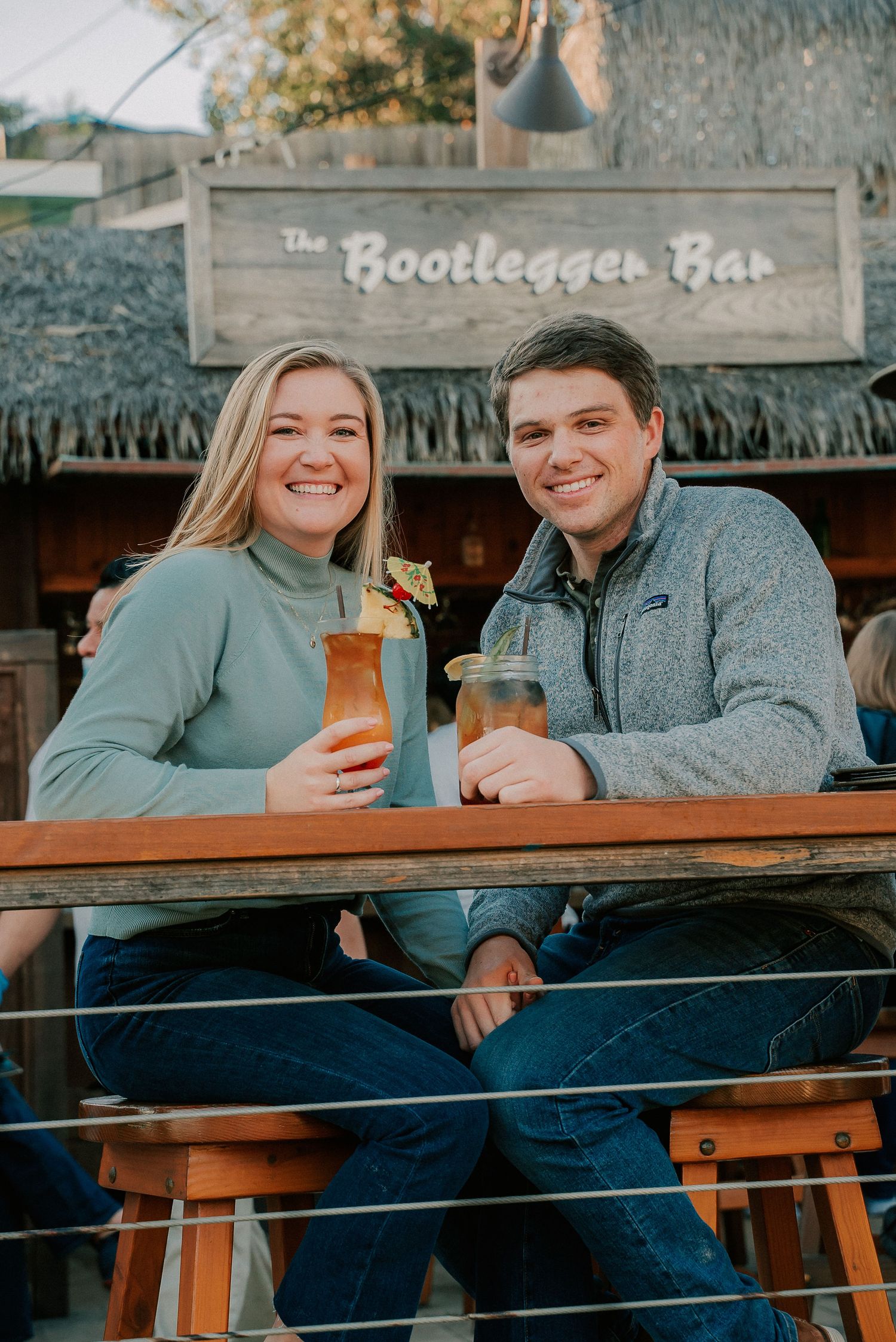 Couple smiling with tropical cocktails at The Bootlegger Bar outdoor patio with tiki-style decor.