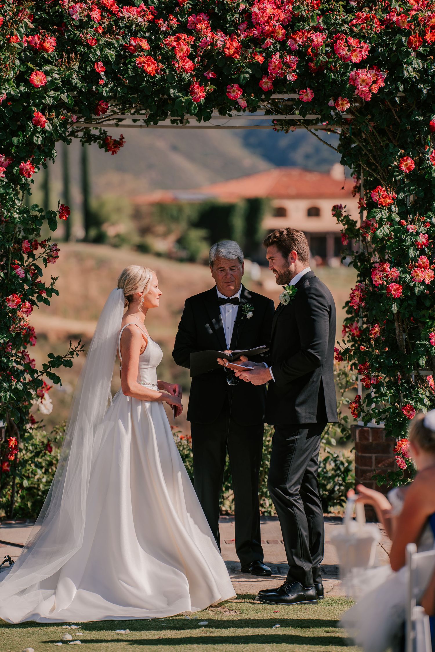 Bride and groom exchange vows under a floral arch adorned with red roses at an outdoor wedding ceremony.