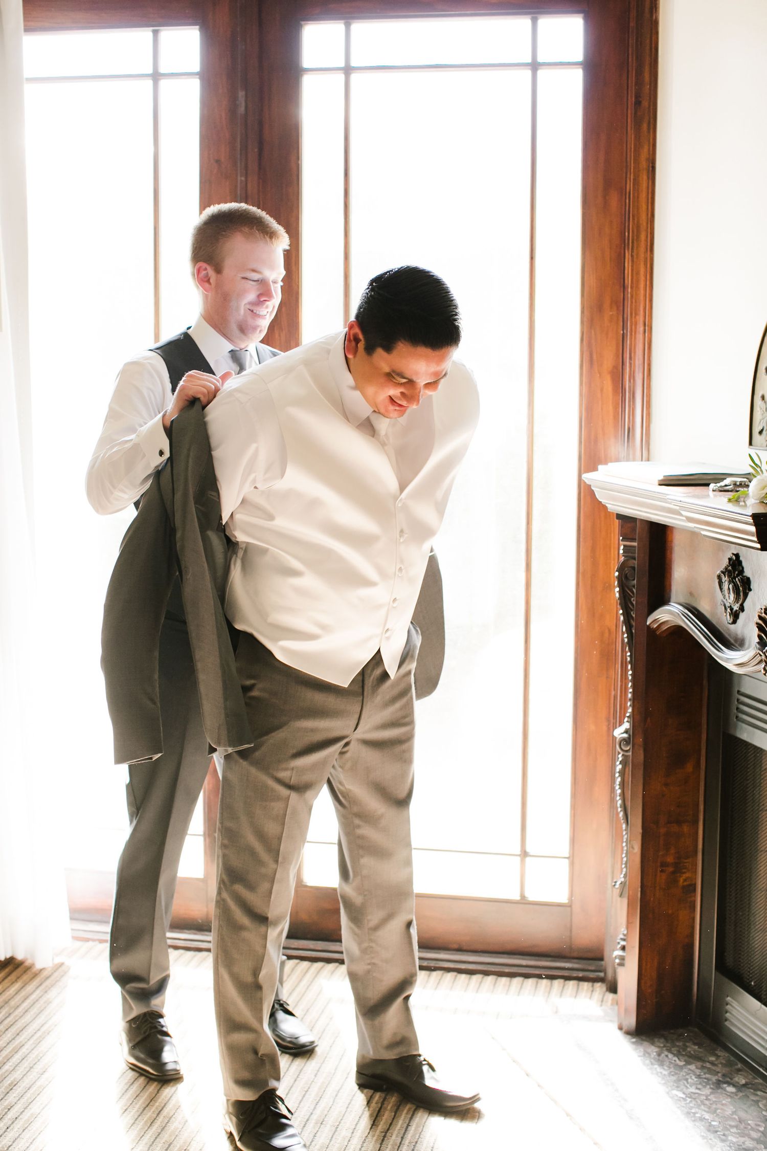 Groomsman helping groom put on suit jacket before wedding ceremony indoors near fireplace.
