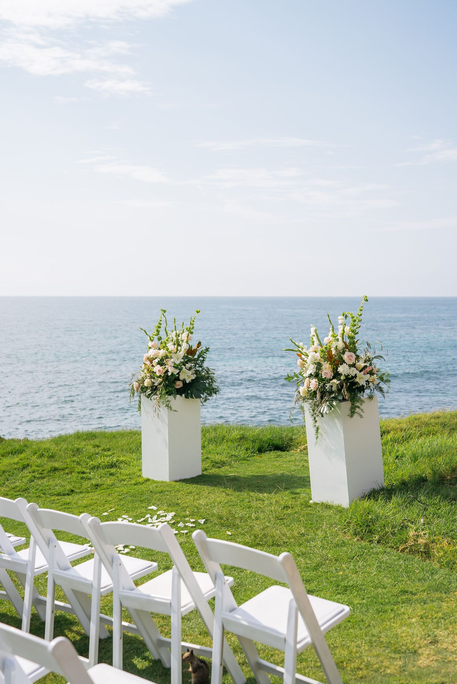 Outdoor oceanside wedding ceremony setup with white chairs and floral arrangements on white pedestals overlooking the sea.