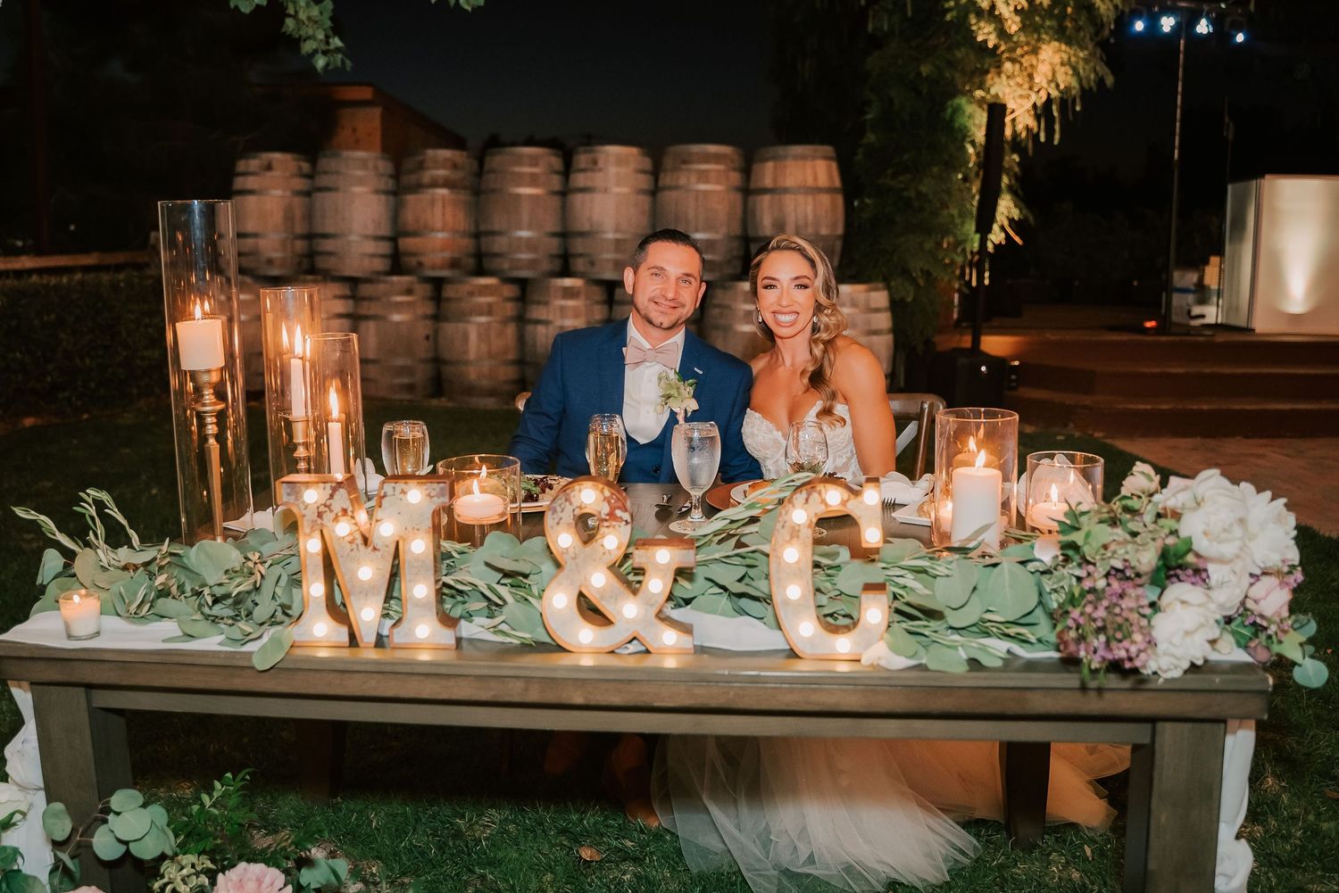 Couple at candlelit wedding sweetheart table with marquee letters, floral greenery, and wine barrels backdrop at night.