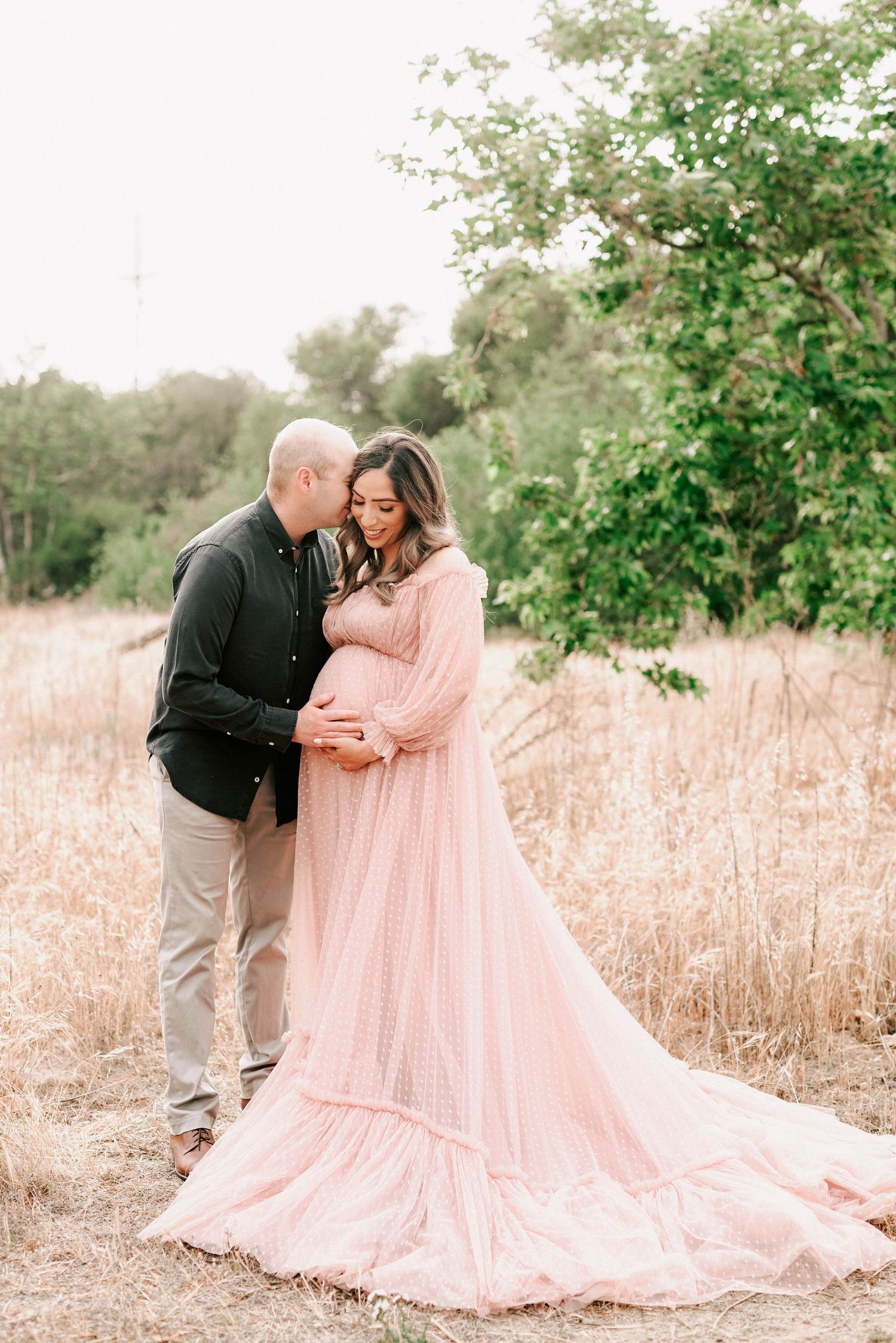 Couple sharing a tender moment in a field, woman in flowing pink maternity gown, surrounded by golden grass and green trees.