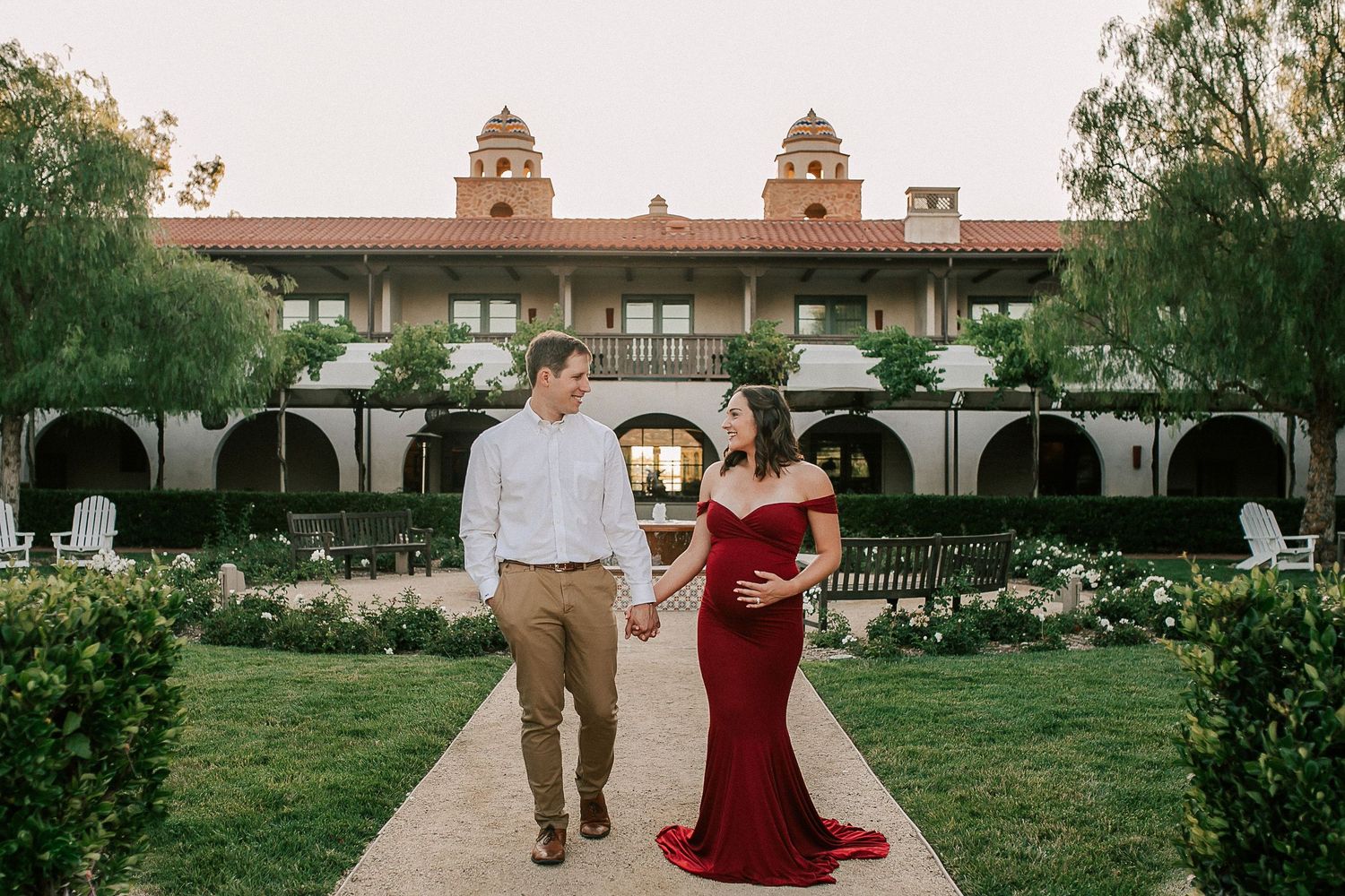 Couple holding hands on garden path, woman in red maternity gown, man in white shirt, elegant Spanish-style building backdrop.