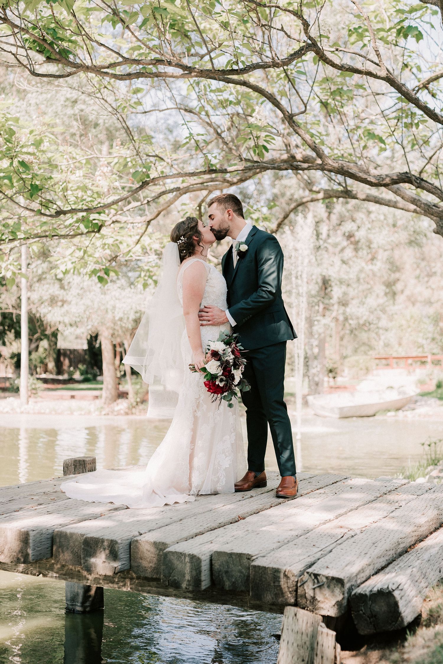 Bride and groom share a romantic kiss on a wooden dock surrounded by lush trees and serene water.