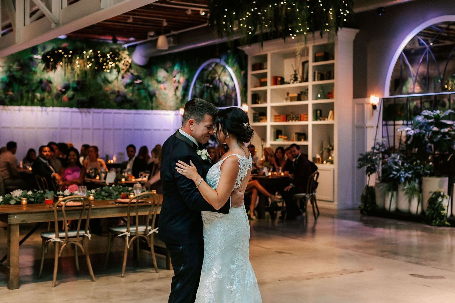 Couple shares their first dance at elegant wedding reception with string lights and lush greenery backdrop.