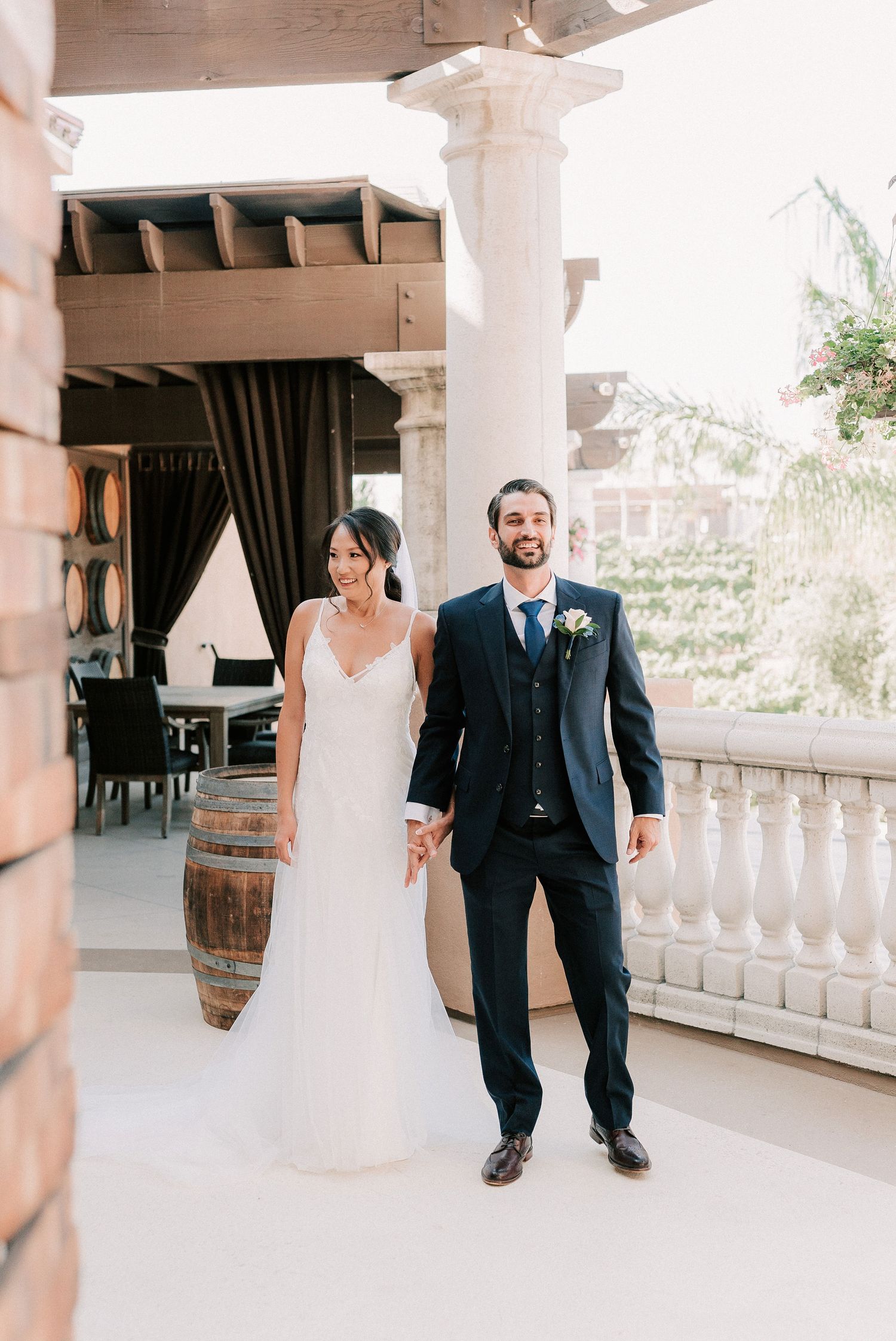 Bride in white gown and groom in navy suit hold hands on elegant venue terrace with stone columns.
