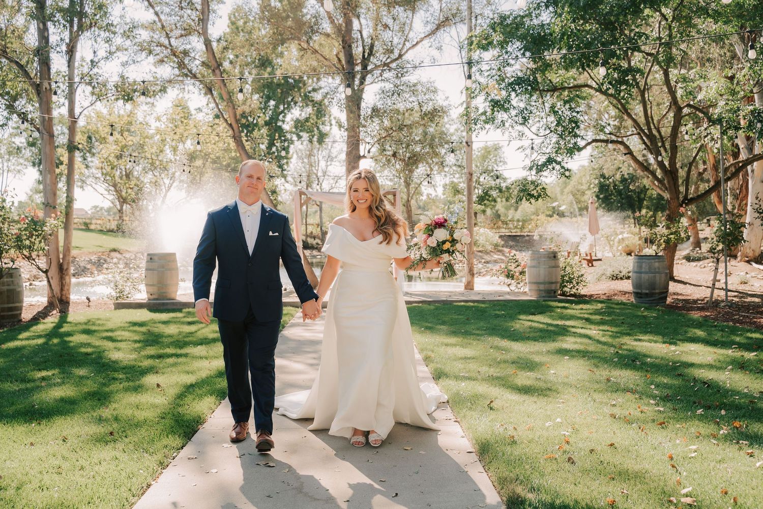 Bride and groom walk hand-in-hand on a garden path at an outdoor wedding venue with lush green trees.