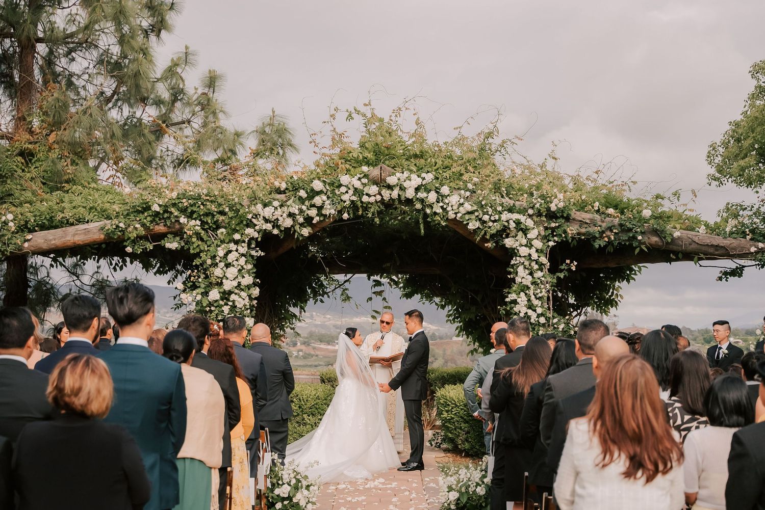 Couple exchanges vows under a floral arch at an outdoor wedding ceremony with guests gathered around.