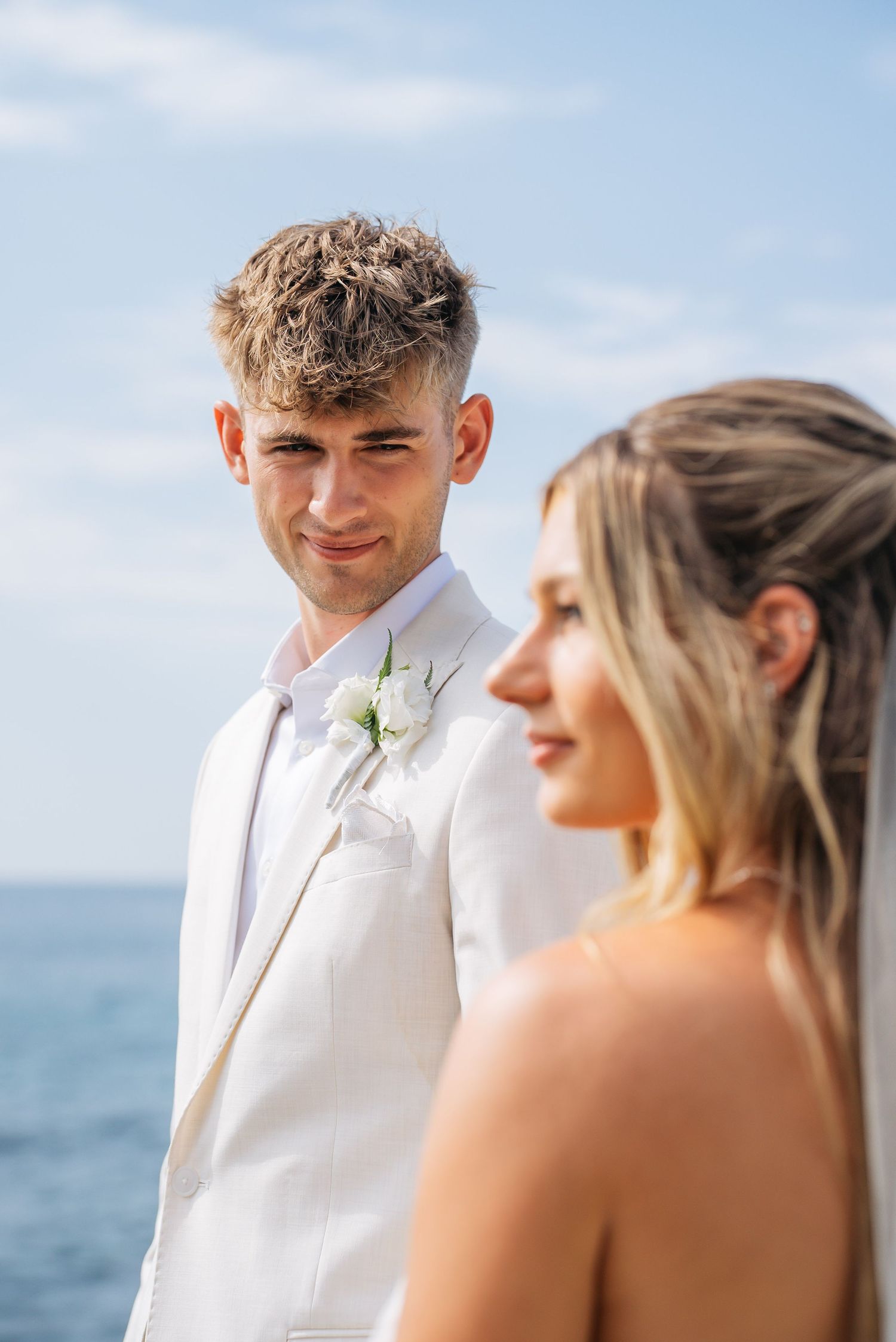 Groom in white suit with boutonniere gazes lovingly at bride with ocean in background.