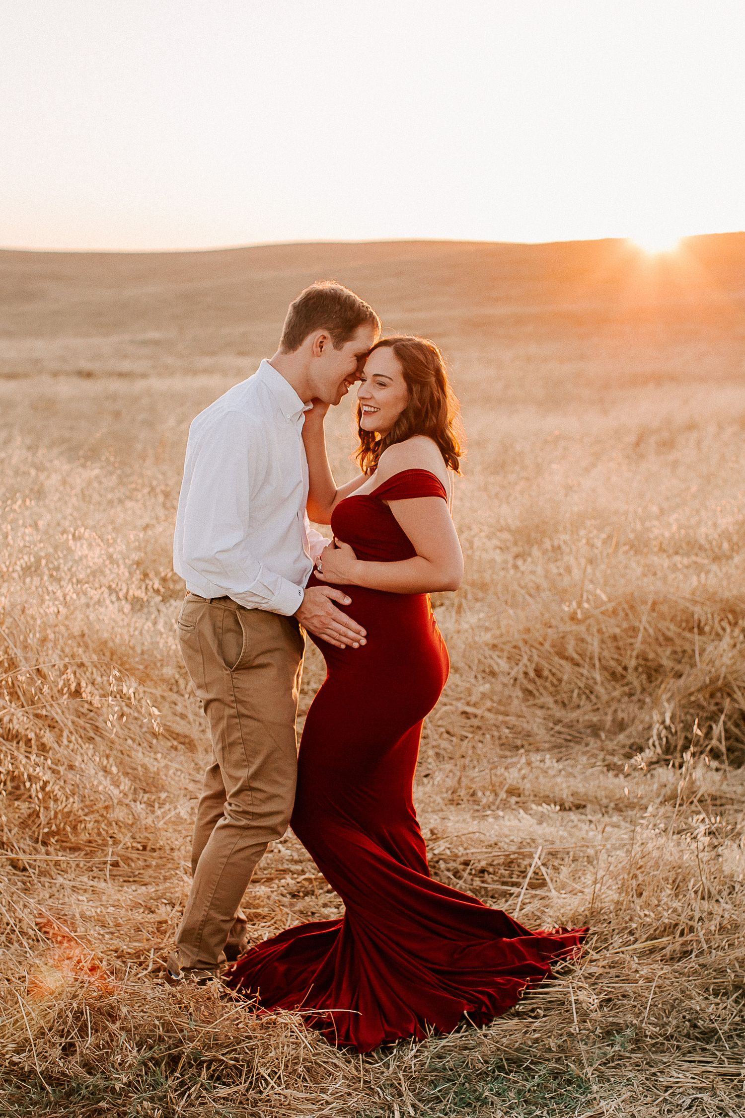Couple poses in golden field at sunset, pregnant woman in red gown, man in white shirt, romantic maternity photo.