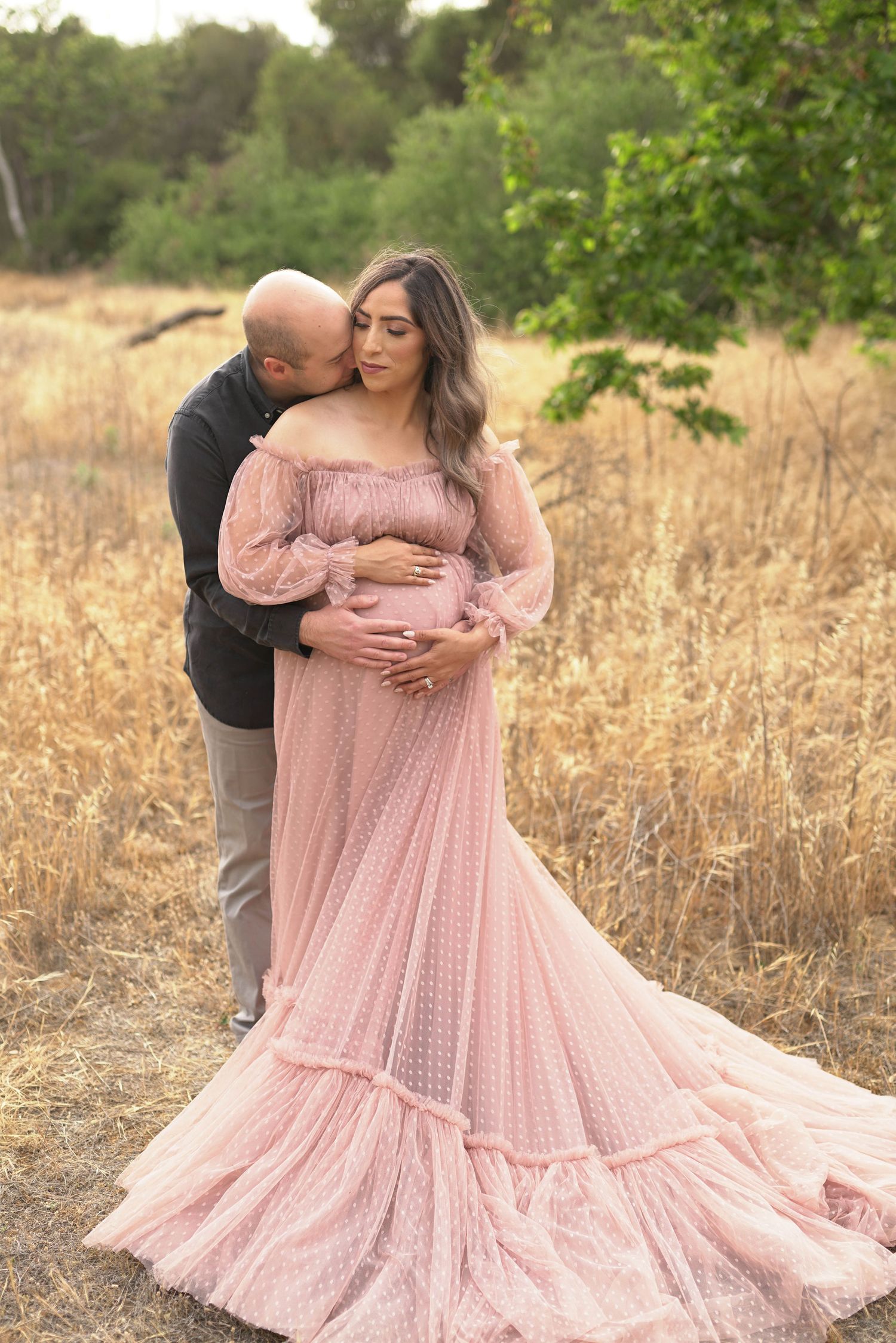 Couple in a golden field, woman in pink maternity gown, man embracing her from behind during outdoor maternity photoshoot.