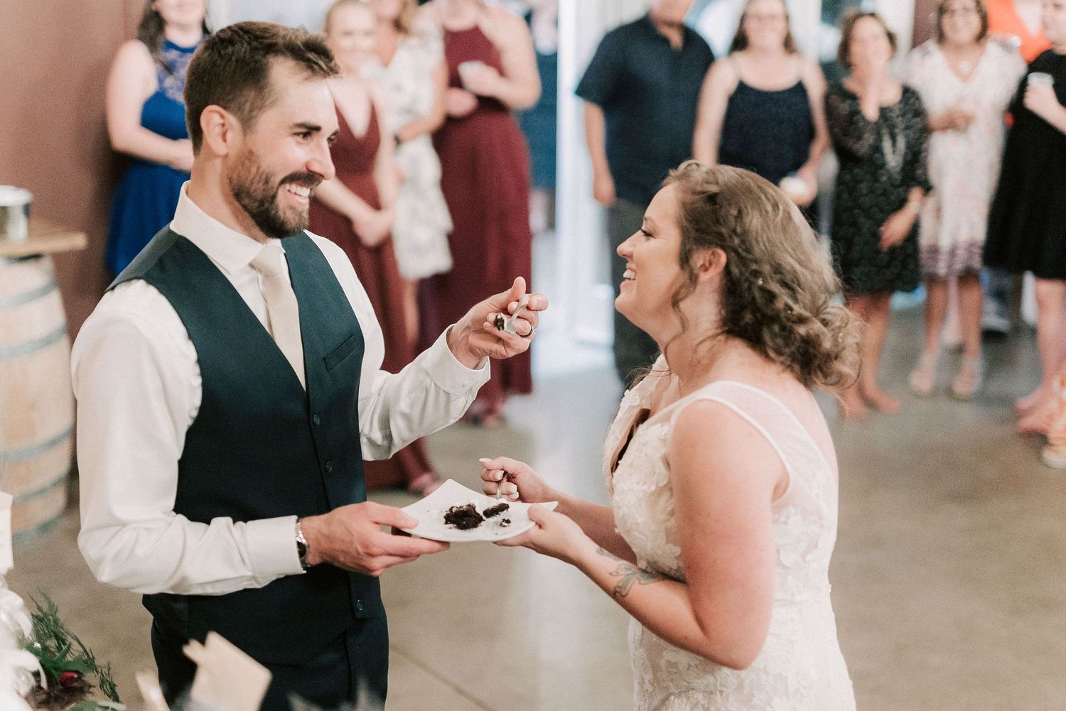 Bride and groom smiling while sharing wedding cake during indoor reception ceremony.