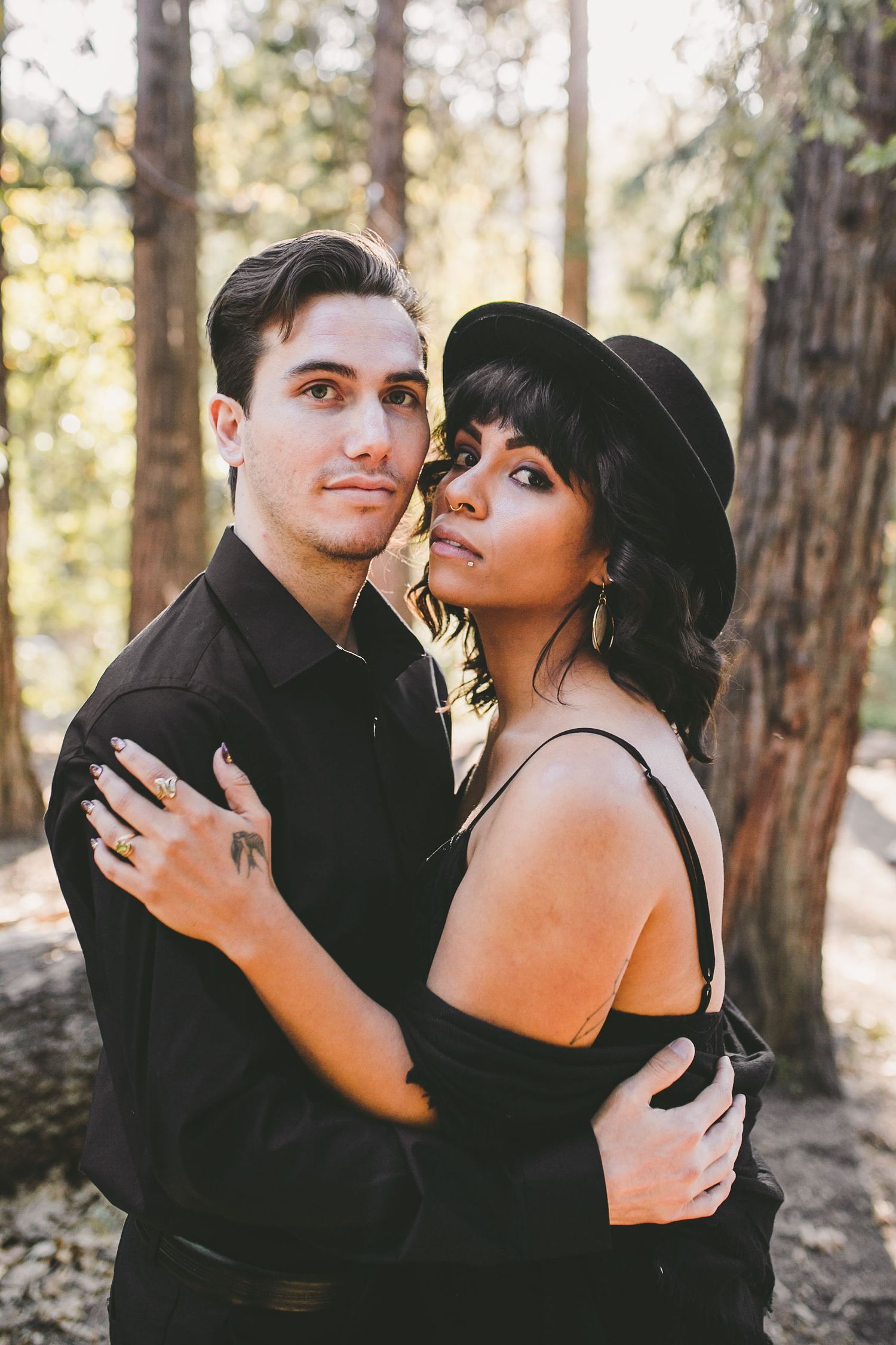 A couple in matching black outfits embraces in a sunlit forest, the woman wearing a black hat.