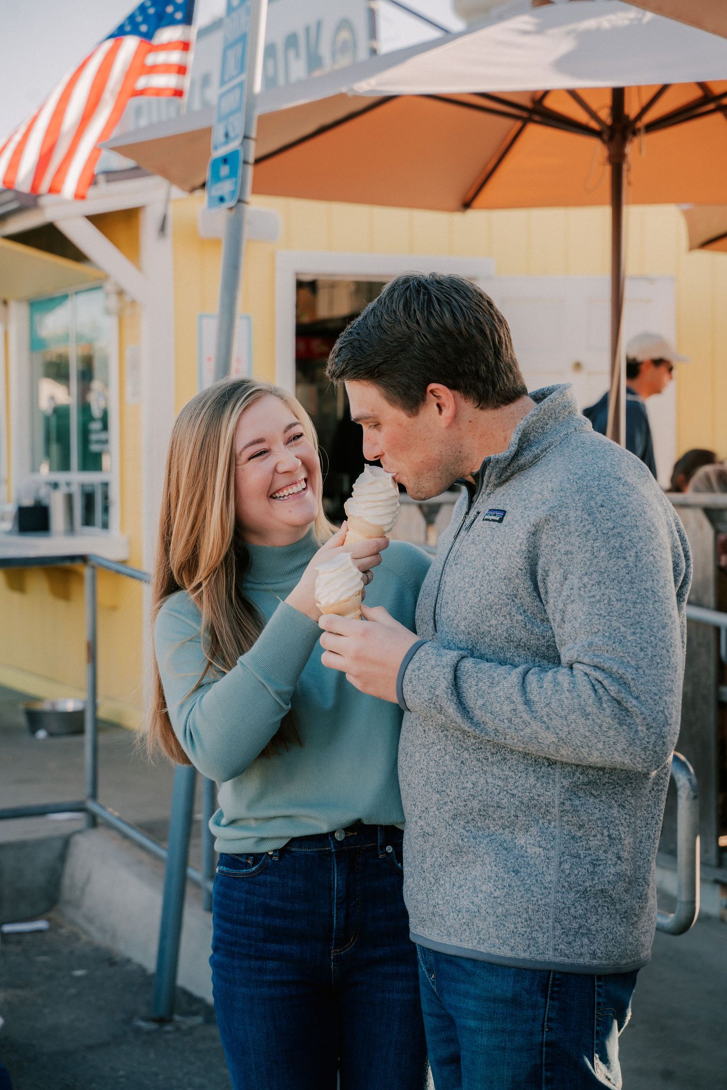 A smiling couple sharing ice cream cones outside a yellow shop with an American flag in the background.