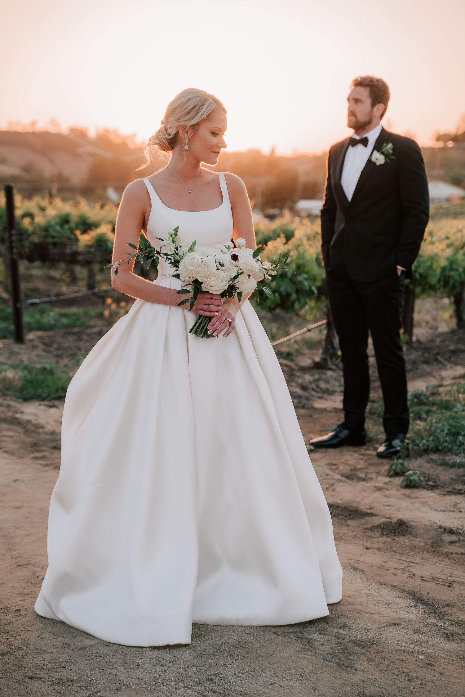 Bride in white ballgown holding bouquet with groom in black tuxedo at sunset vineyard wedding.