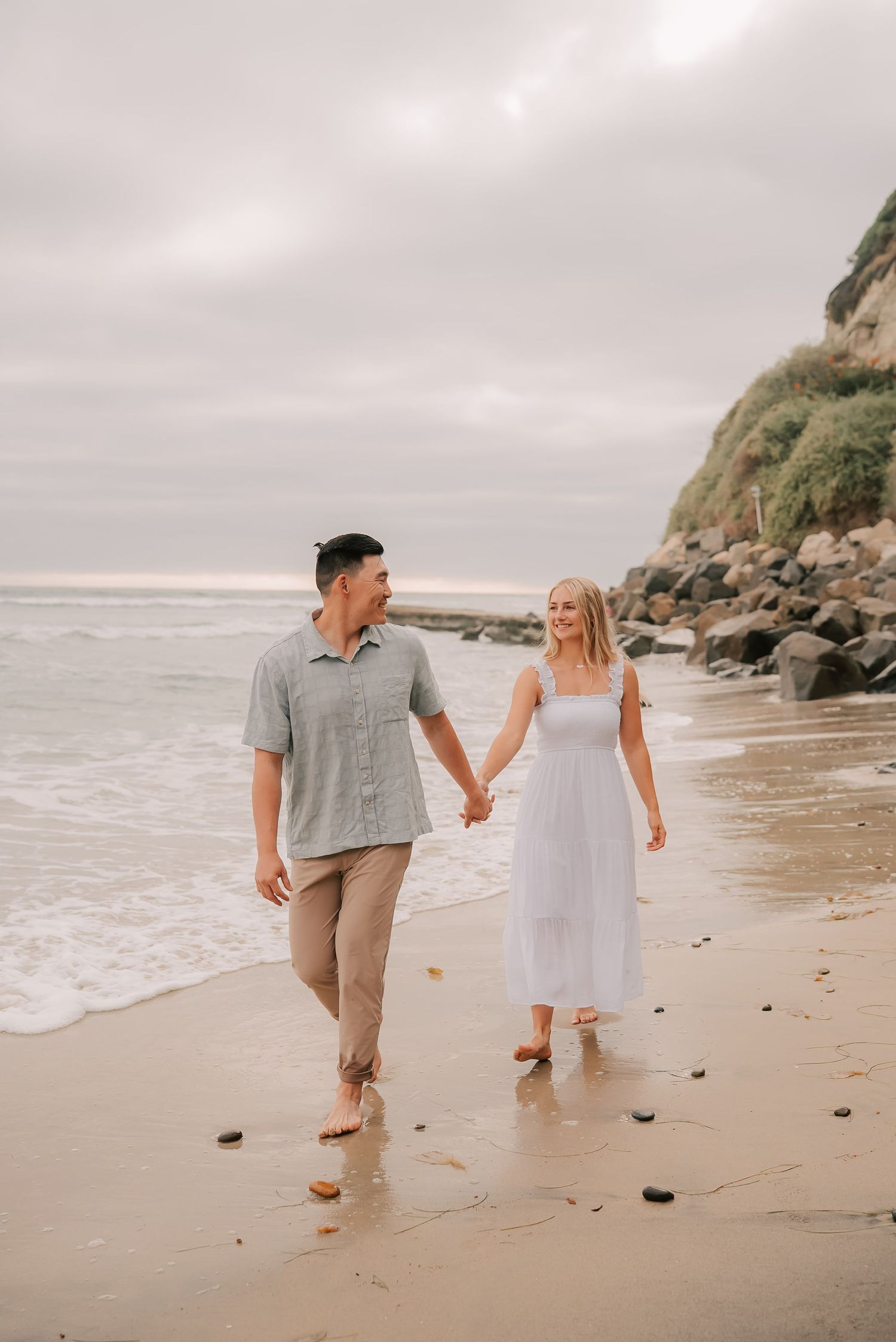 A couple walks hand-in-hand along a sandy beach, the woman in a white dress and man in a gray shirt, with ocean waves nearby.
