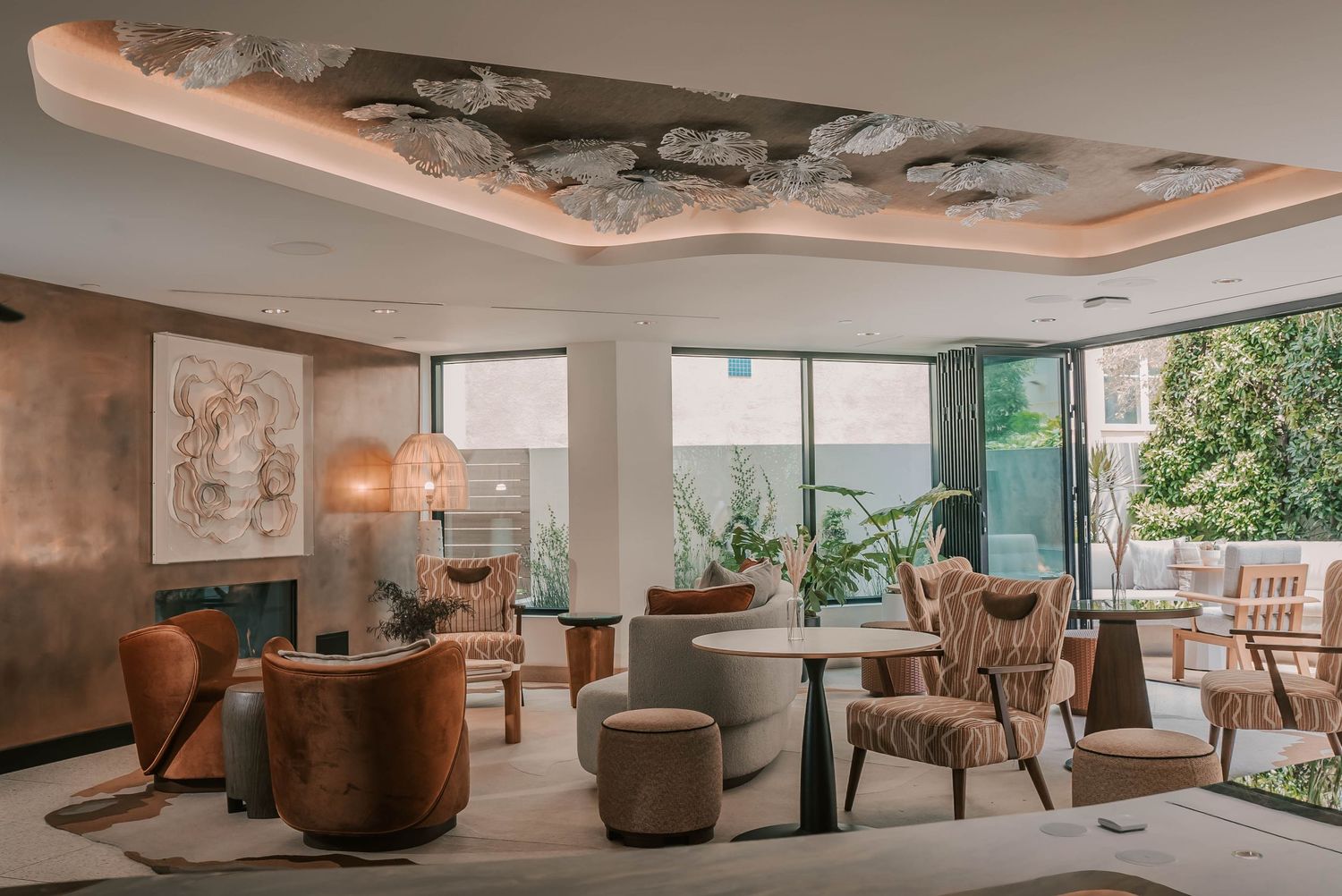 Elegant hotel lounge with velvet armchairs, marble tables, decorative ceiling, and floor-to-ceiling windows overlooking a garden.