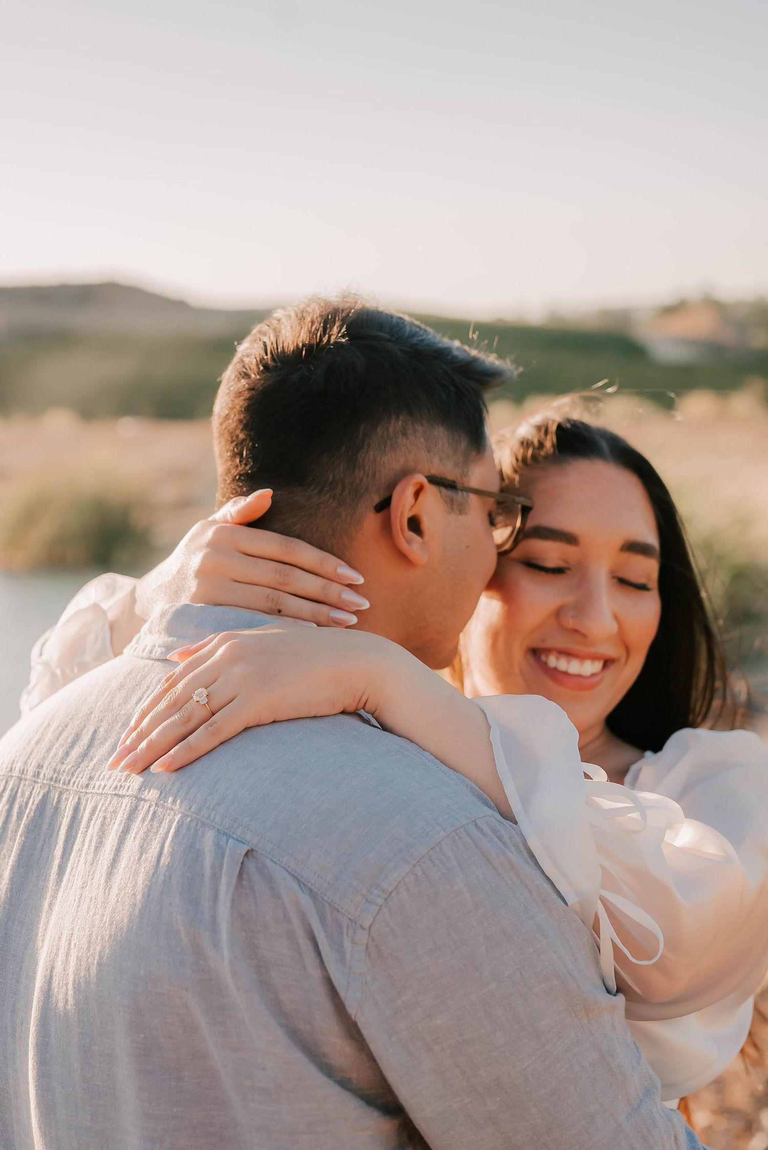 Couple embracing outdoors at golden hour, woman smiling with engagement ring visible, soft natural background.