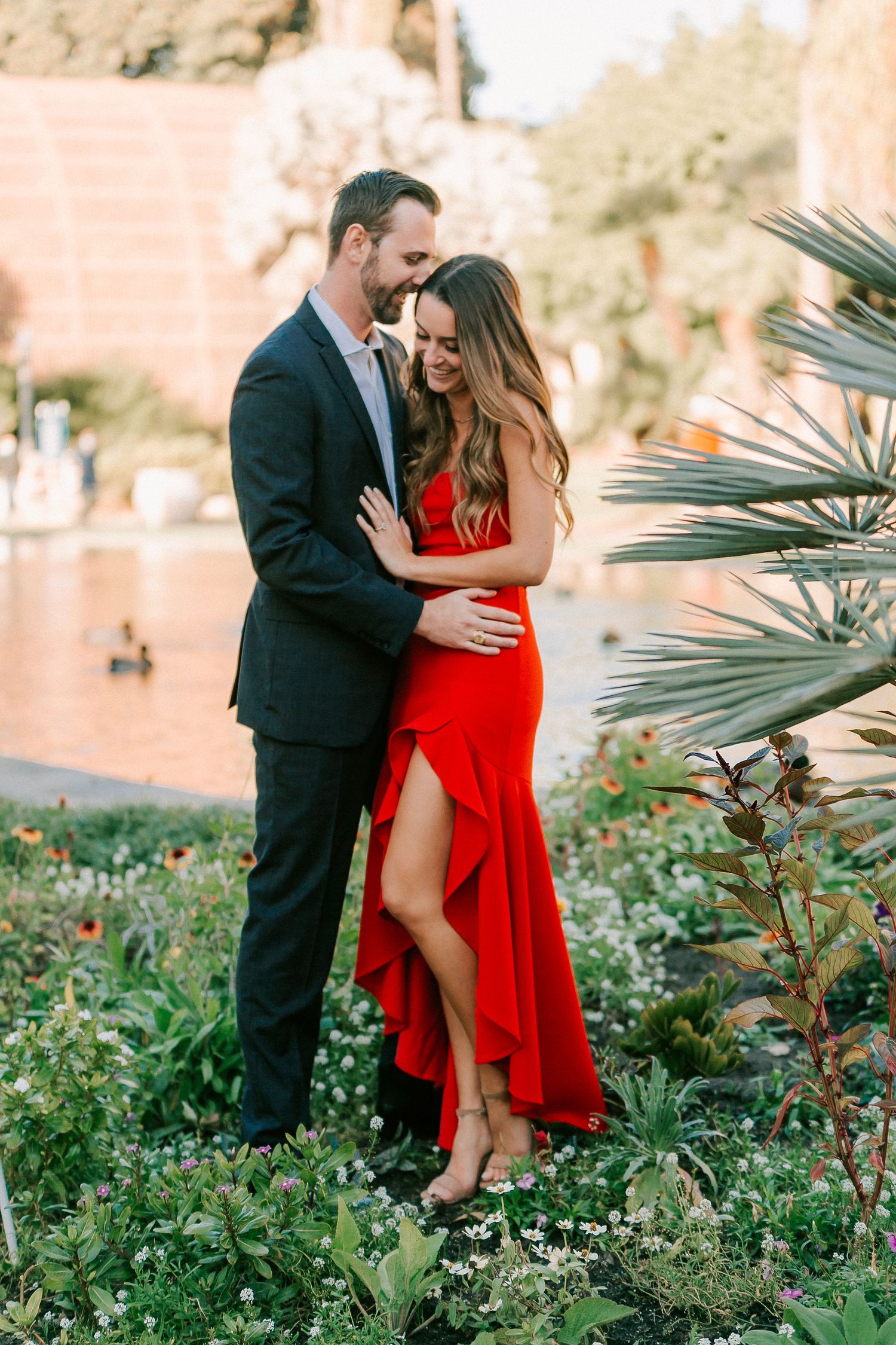 Couple embracing in a lush garden, woman in elegant red dress with a ruffled slit skirt.