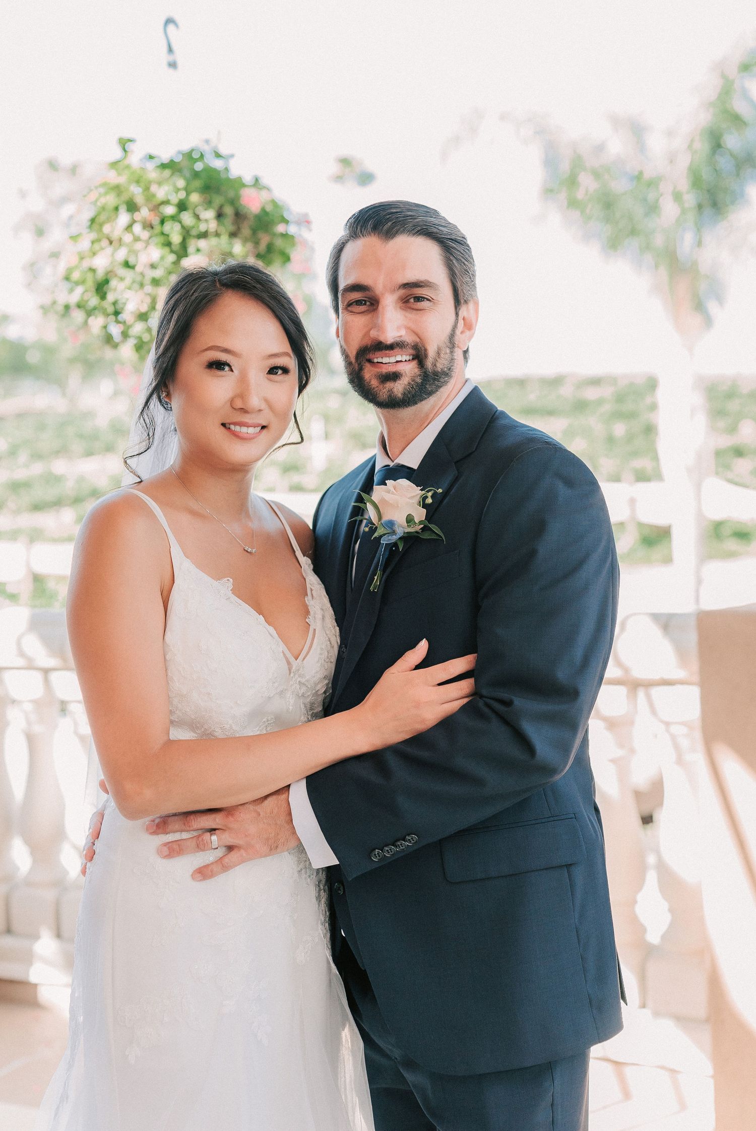 Smiling bride in white lace gown and groom in navy suit with boutonniere pose together at outdoor wedding venue.